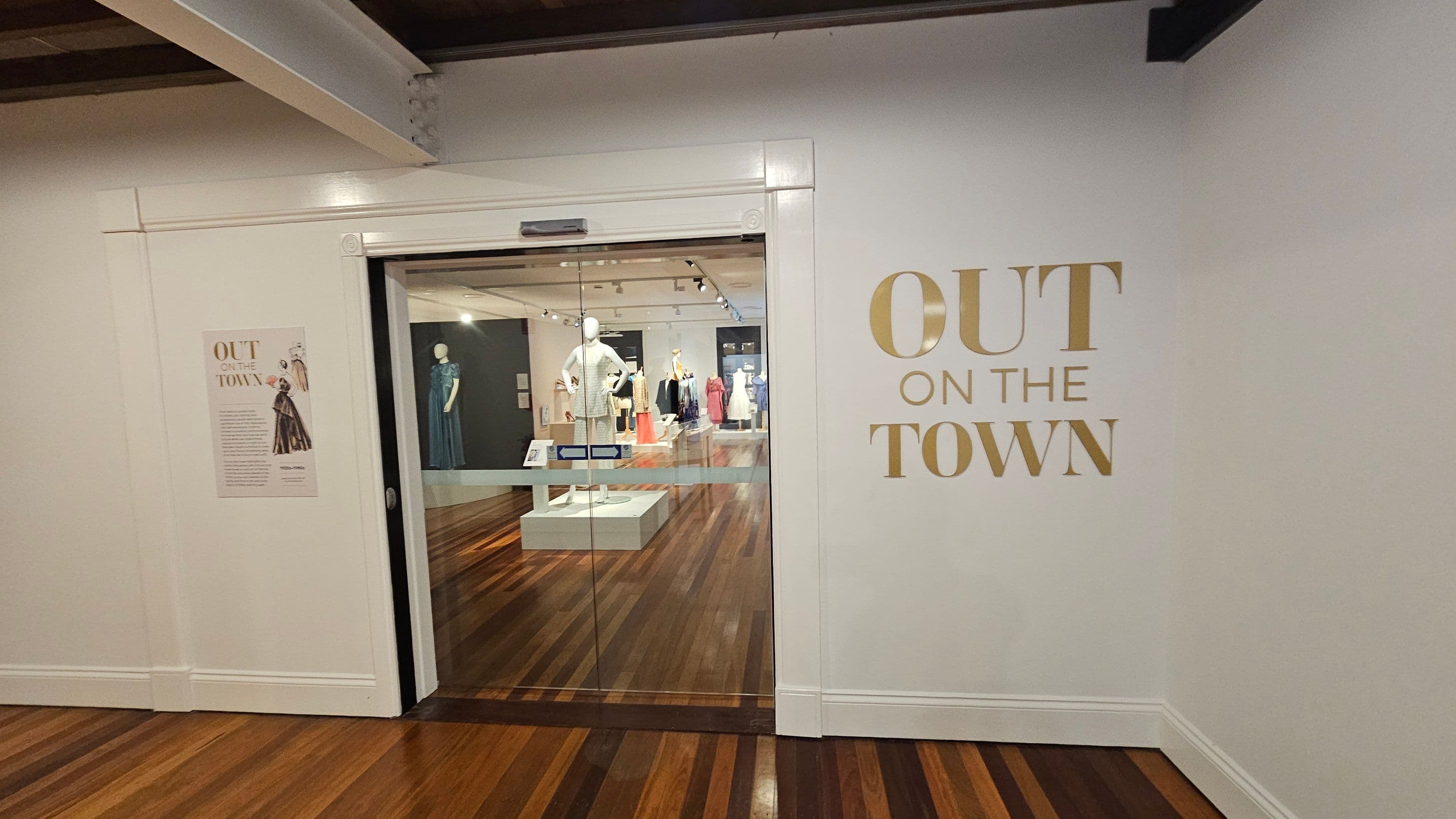 A museum entrance with a glass door in the centre, leading to an exhibition space. On the right wall, large gold letters spell "OUT ON THE TOWN". To the left, a poster with text and an illustration is mounted on the wall. Inside the exhibition, mannequins display various outfits on platforms. The floor is wooden, and the ceiling has exposed beams and track lighting.