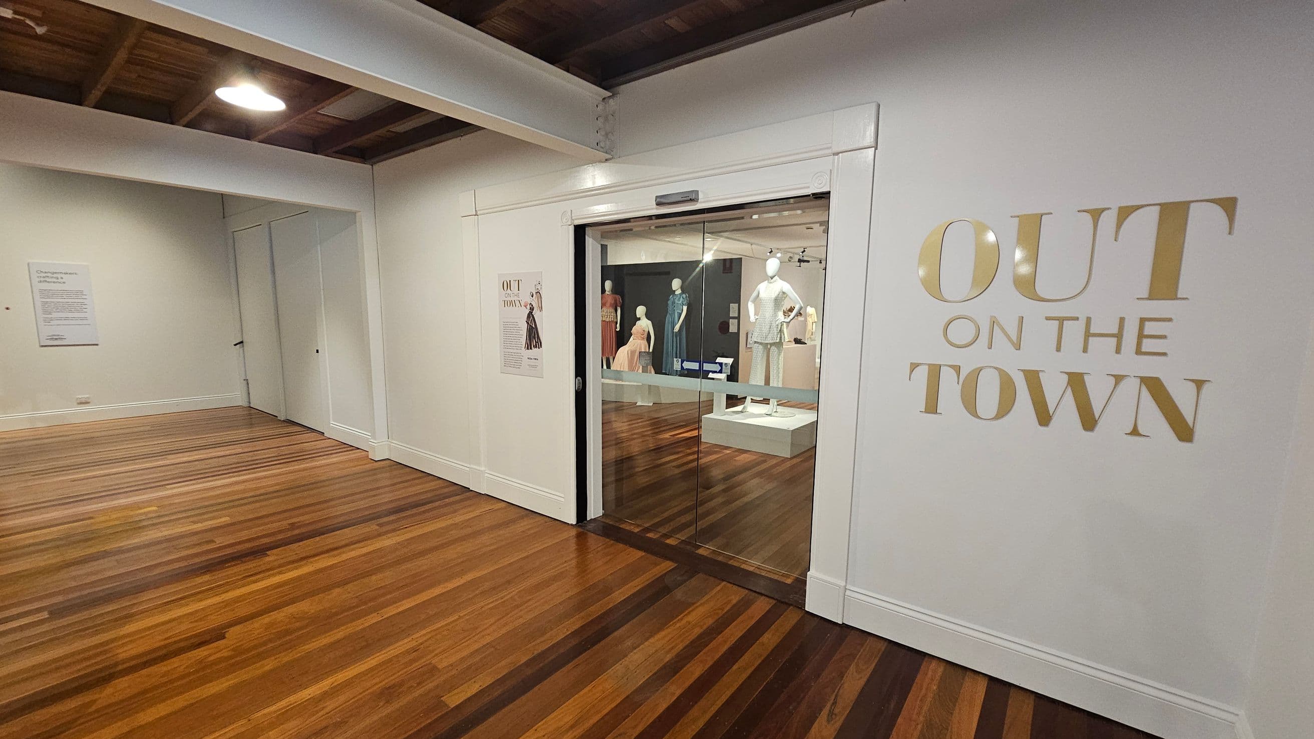 A hallway with wooden flooring and a white ceiling with exposed beams. On the right, a wall displays the words "OUT ON THE TOWN" in large gold letters. Below, a glass door leads to a room with mannequins dressed in various outfits. On the left, a wall features a sign with text and a closed door. The lighting is bright, with a ceiling light visible.