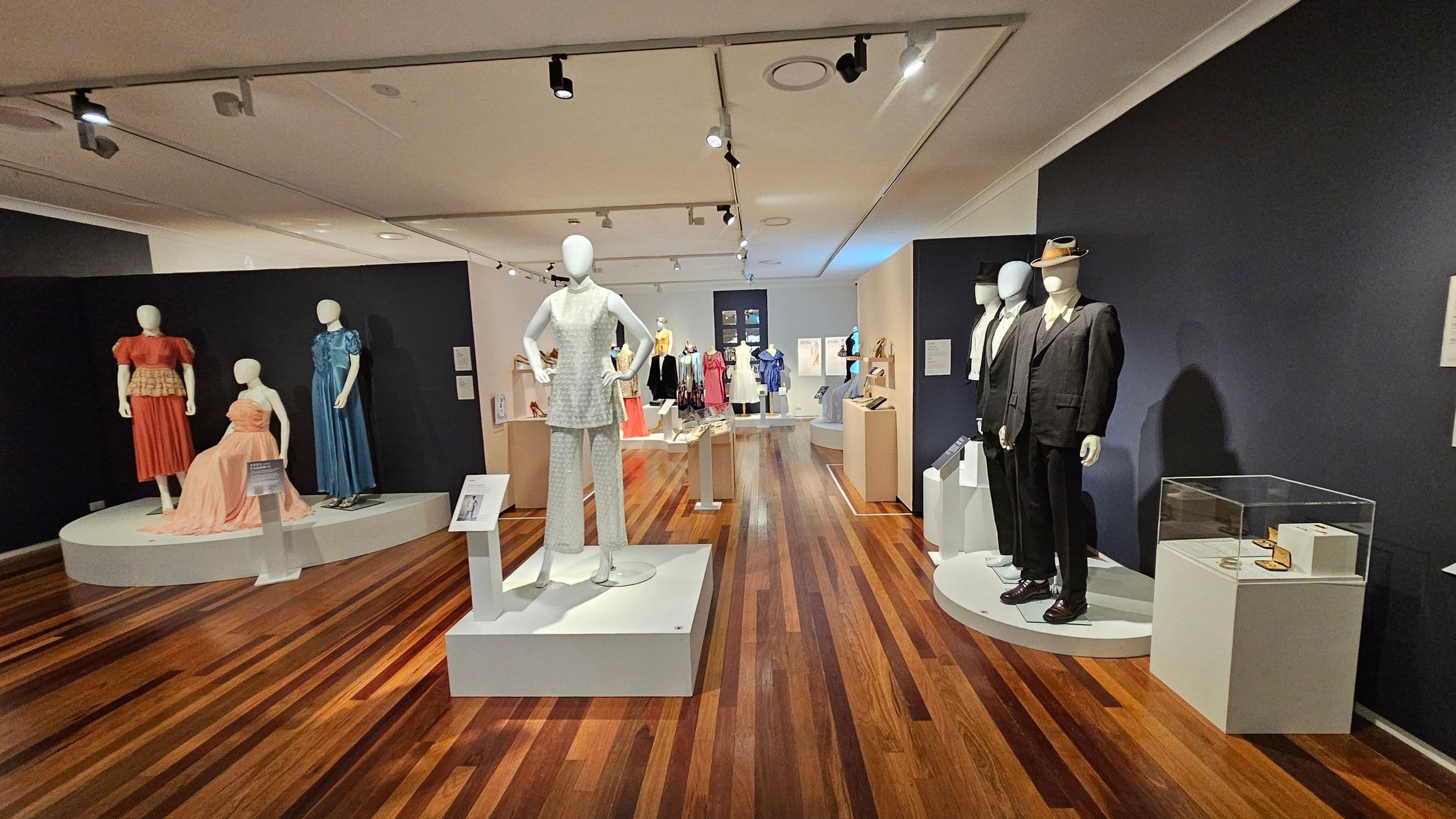 A museum exhibit featuring mannequins dressed in vintage clothing. On the left, three mannequins display dresses in red, peach, and blue on a circular platform. In the centre, a mannequin wears a white outfit on a square pedestal. On the right, two mannequins in dark suits and hats stand on a round platform. The background shows more mannequins in various outfits. The floor is wooden, and the ceiling has track lighting.
