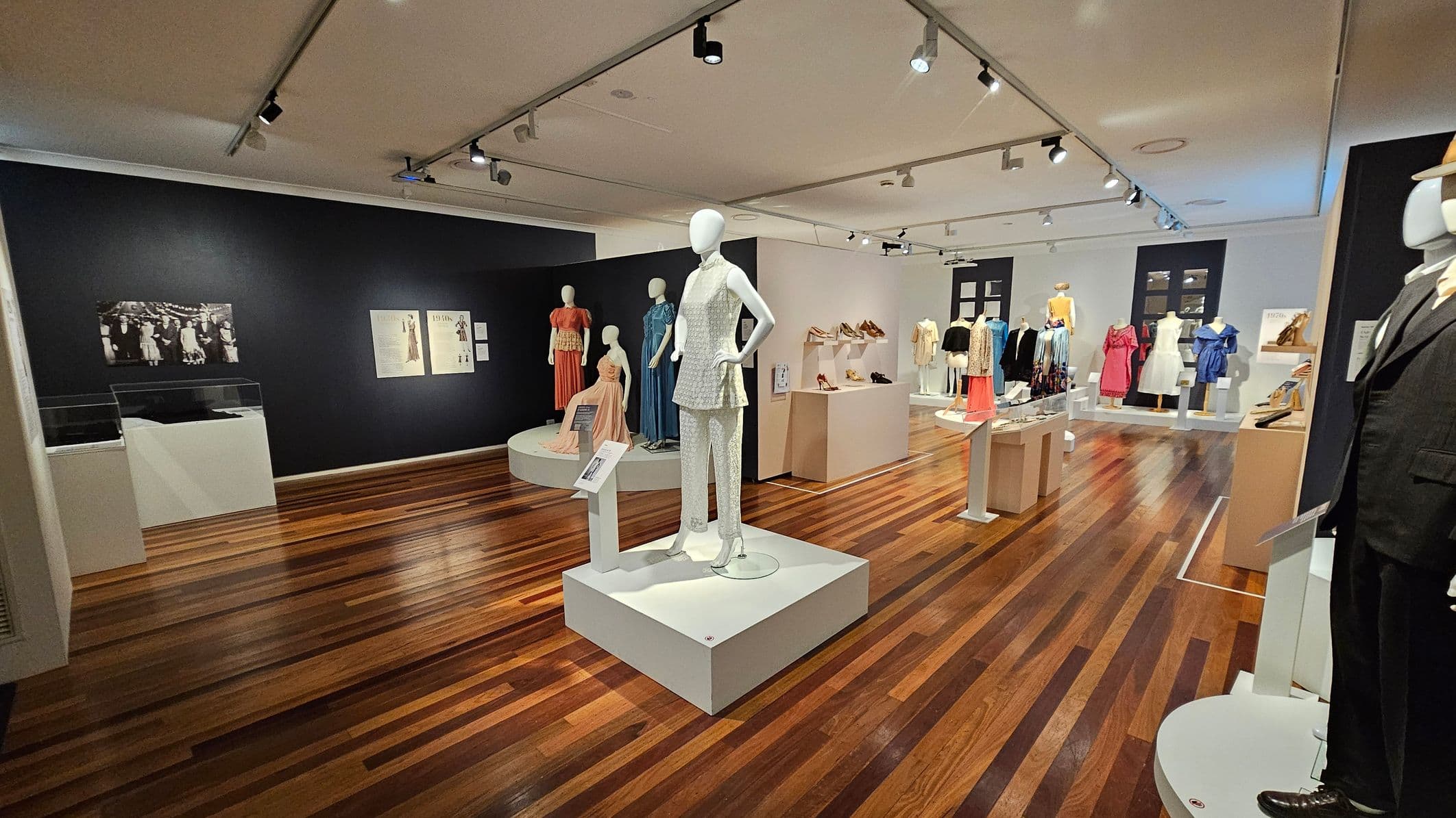 A fashion exhibition room with wooden flooring and a white ceiling featuring track lighting. In the centre, a mannequin displays a white outfit on a raised platform. To the left, mannequins in colourful dresses are positioned against a black wall with framed images and text. On the right, more mannequins showcase various outfits, with shelves displaying shoes. A mannequin in a dark suit is partially visible in the foreground on the right.