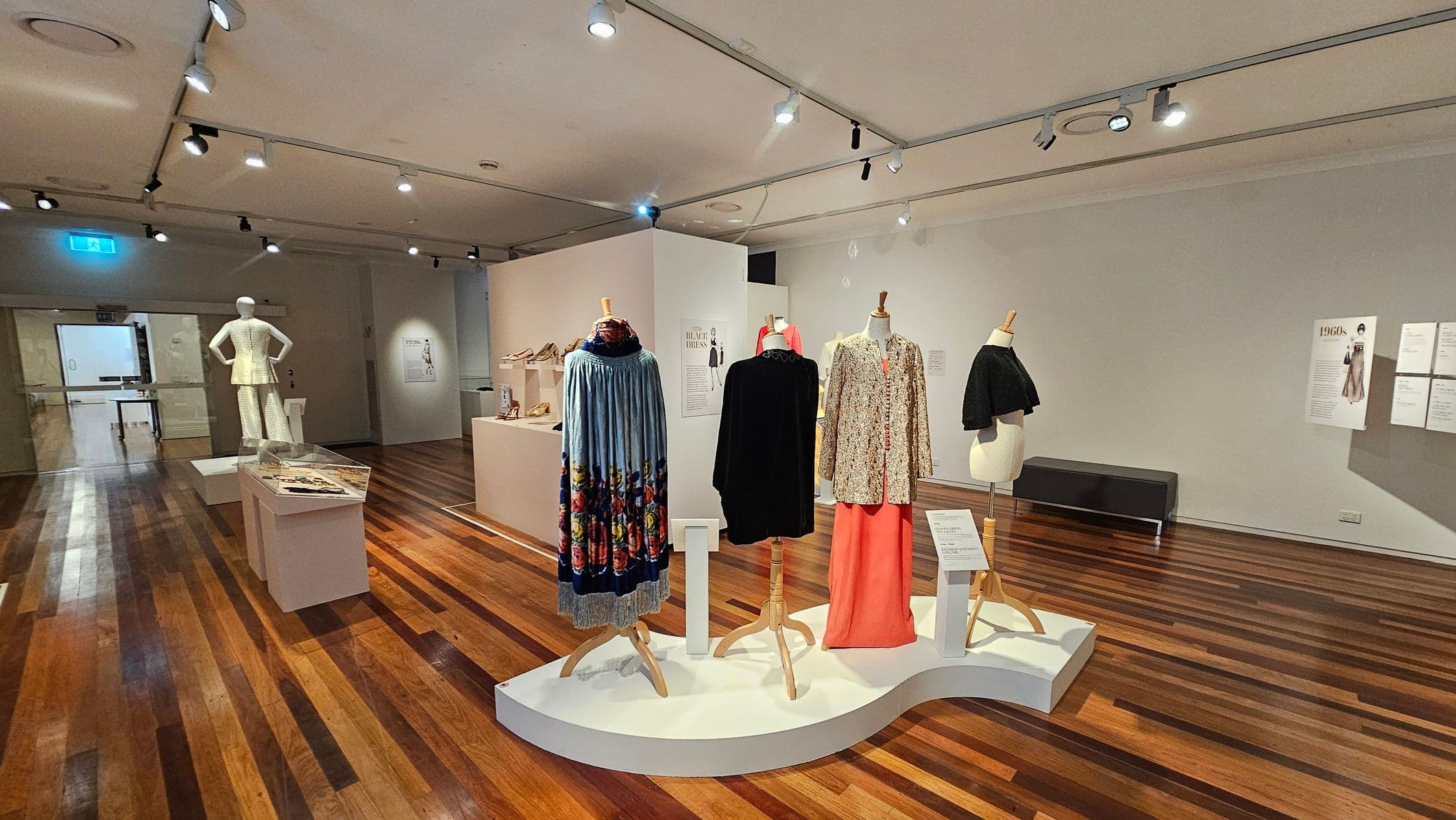 A museum exhibit with mannequins displaying vintage clothing. In the centre, three mannequins showcase garments including a blue floral dress, a black coat, and a patterned coat with a pink skirt. To the left, a mannequin in a white outfit stands near a display case with accessories. The floor is wooden, and the ceiling has track lighting. On the right, informational posters are mounted on the wall. A bench is positioned against the wall in the background.