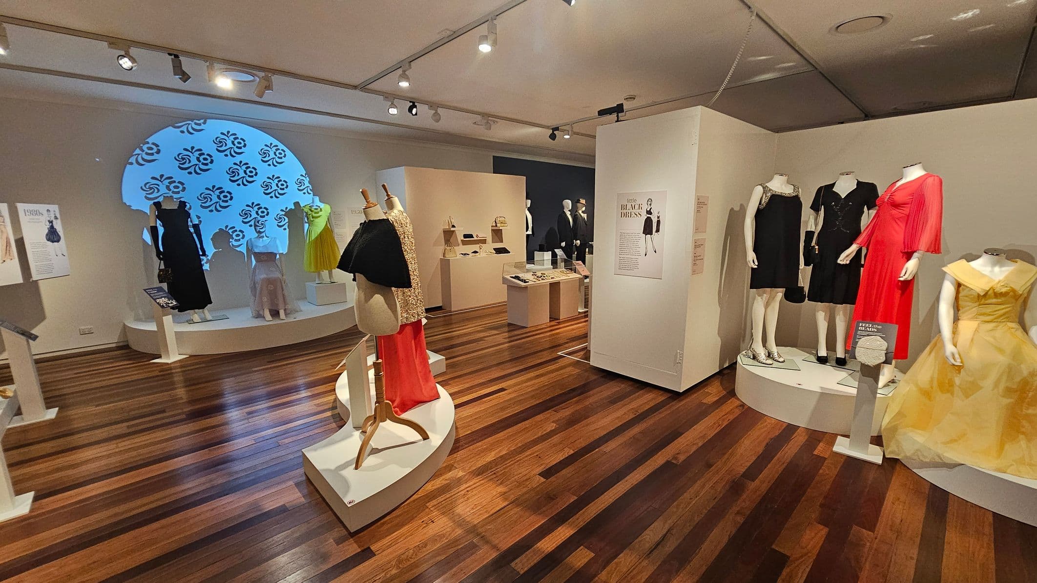 A fashion exhibit with mannequins displaying various dresses. On the left, a black dress and a yellow dress are showcased against a wall with a decorative blue pattern. In the centre, a mannequin wears a red skirt and a black top. On the right, three mannequins display a red dress, a black dress, and a yellow gown. The floor is wooden with a polished finish, and the ceiling has track lighting. Information panels and display cases are scattered throughout the space.