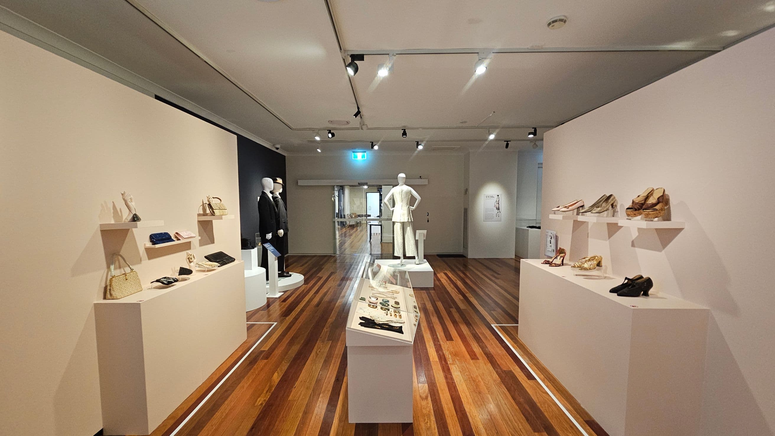 A museum exhibit with wooden flooring and a white ceiling. On the left, shelves display handbags and accessories. In the centre, a mannequin in a white outfit stands on a platform, with a glass case of jewellery and gloves in front. On the right, shelves showcase various shoes. In the background, two mannequins in dark suits are visible, and an exit sign is above a doorway leading to another room. Track lighting illuminates the space.