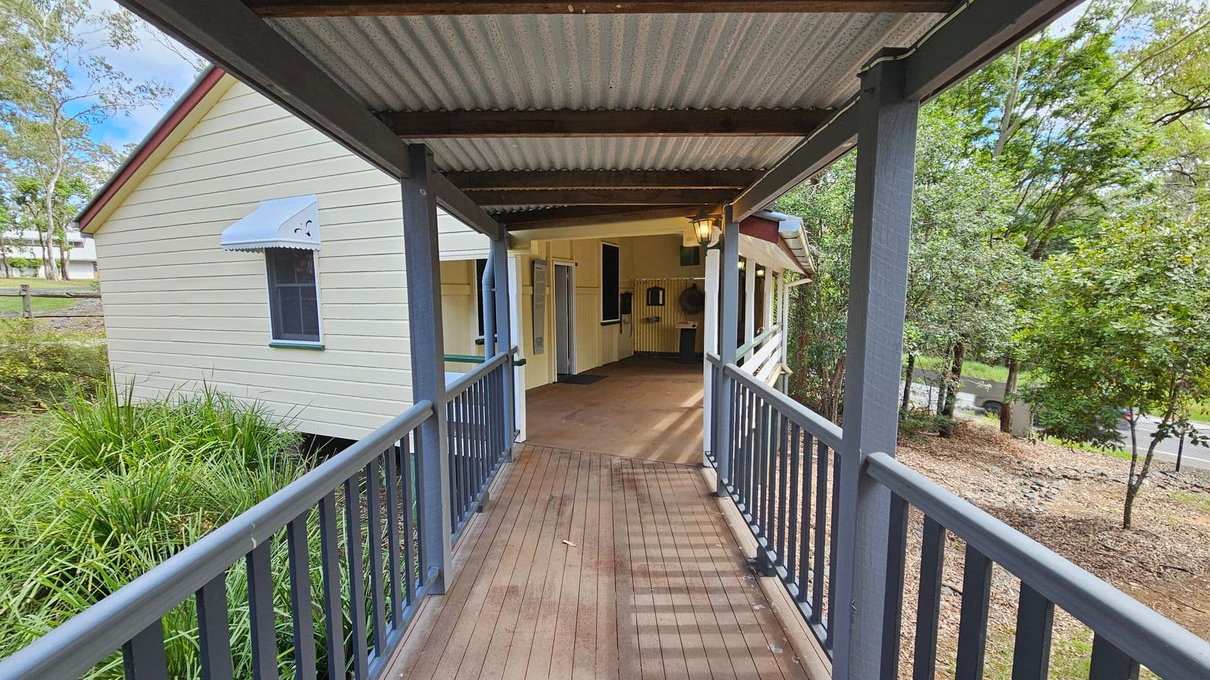 A covered wooden walkway with a corrugated metal roof leads to a building. On the left, there's a cream-coloured wall with a small window featuring a blue awning. The right side has a railing and overlooks a garden area with trees and shrubs. The floor is wooden, and the ceiling is made of corrugated metal. In the background, the entrance to the building is visible, with a light fixture hanging above.