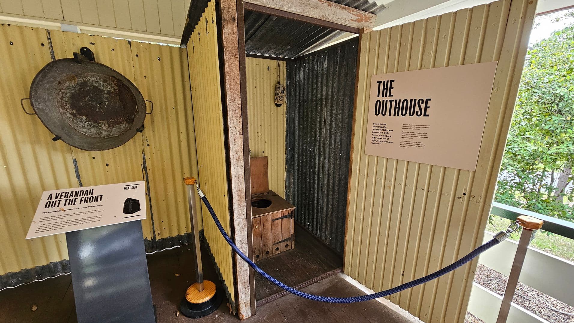 An indoor exhibit featuring a small outhouse structure made of corrugated metal. On the left, a large, round metal object is mounted on the wall above a sign titled "A Verandah Out the Front." The outhouse, in the centre, has a wooden seat with a hole. On the right, a sign titled "The Outhouse" provides information. A blue rope barrier is in the foreground, and greenery is visible through a window on the right. The floor is concrete, and the ceiling is corrugated metal.