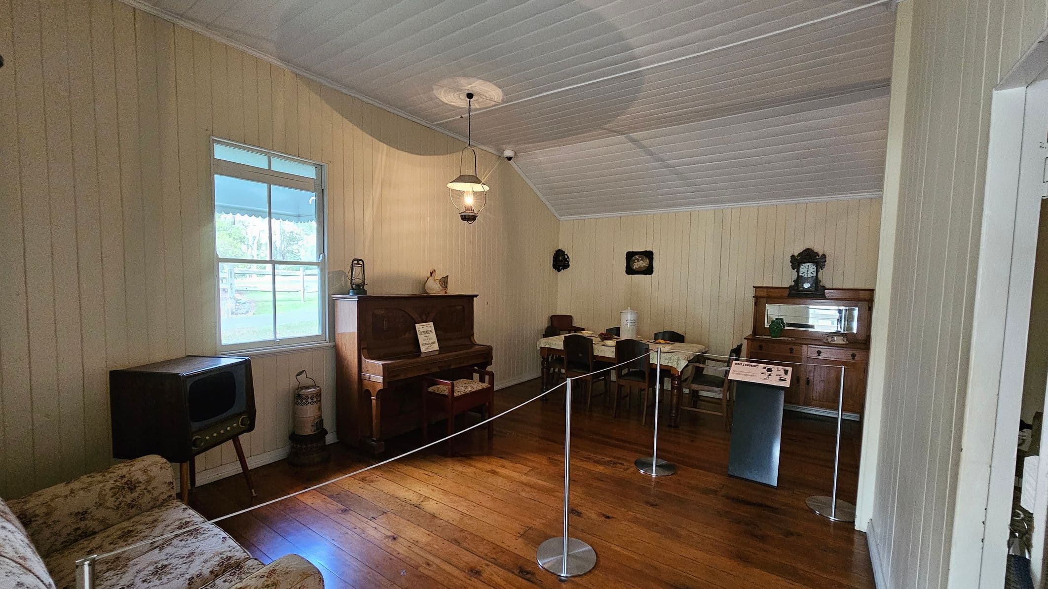A vintage room with wooden floors and a white panelled ceiling. On the left, there's a floral-patterned sofa and an old television set. A window with a view of a garden is above the TV. In the centre, a wooden piano with a small chair is placed against the wall, with a lantern and decorative items on top. On the right, a dining table with chairs and a wooden sideboard with a clock and mirror. A rope barrier separates the room from the viewer.