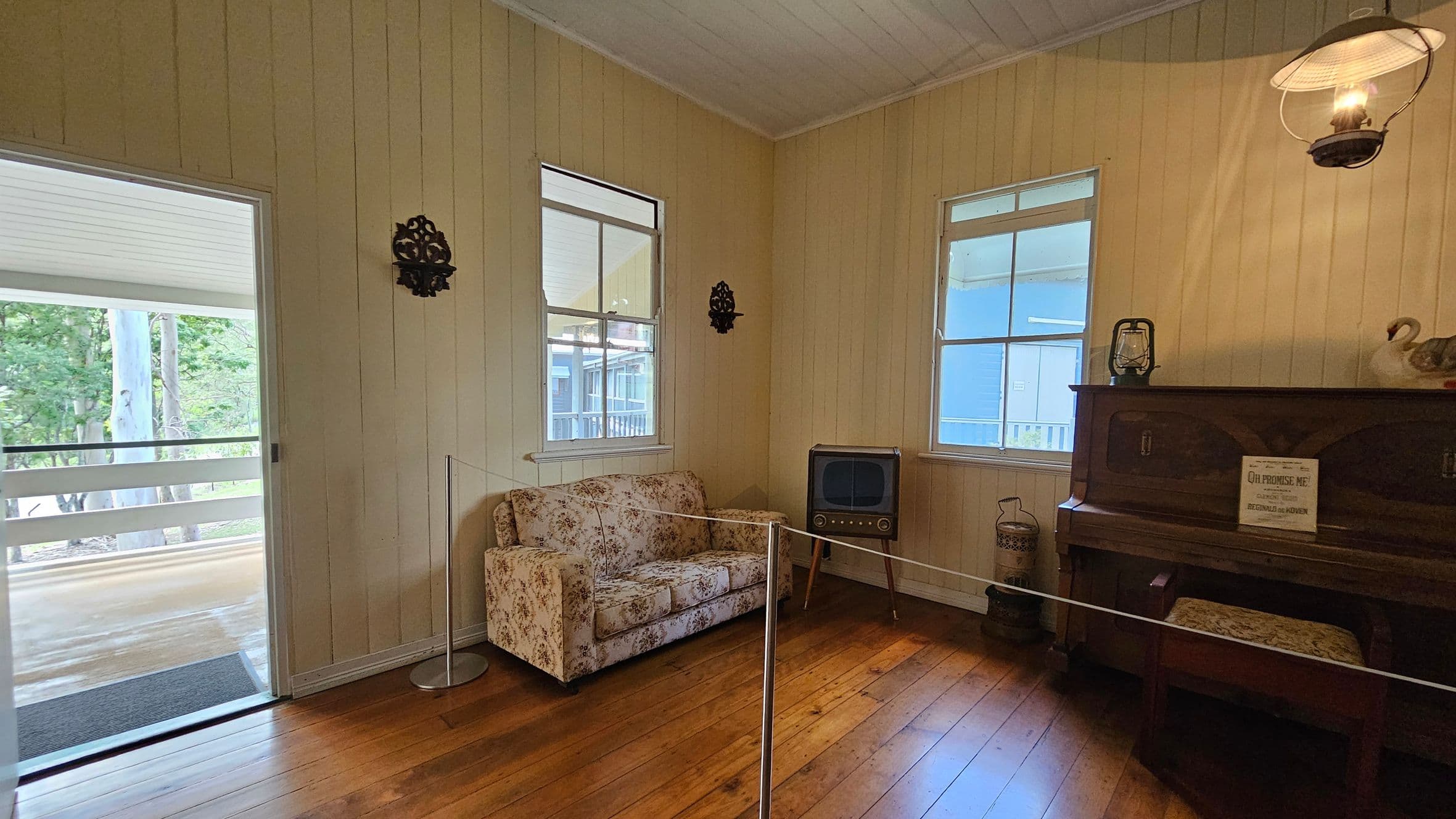 A room with wooden panelled walls and a wooden floor. On the left, there's an open door leading to a porch with a view of trees. Inside, a floral-patterned sofa sits against the wall beneath two windows. In the corner, an old-fashioned television on legs. On the right, a wooden piano with a small sign on top, alongside a decorative swan and a lantern. A wall-mounted lamp provides light above the piano.