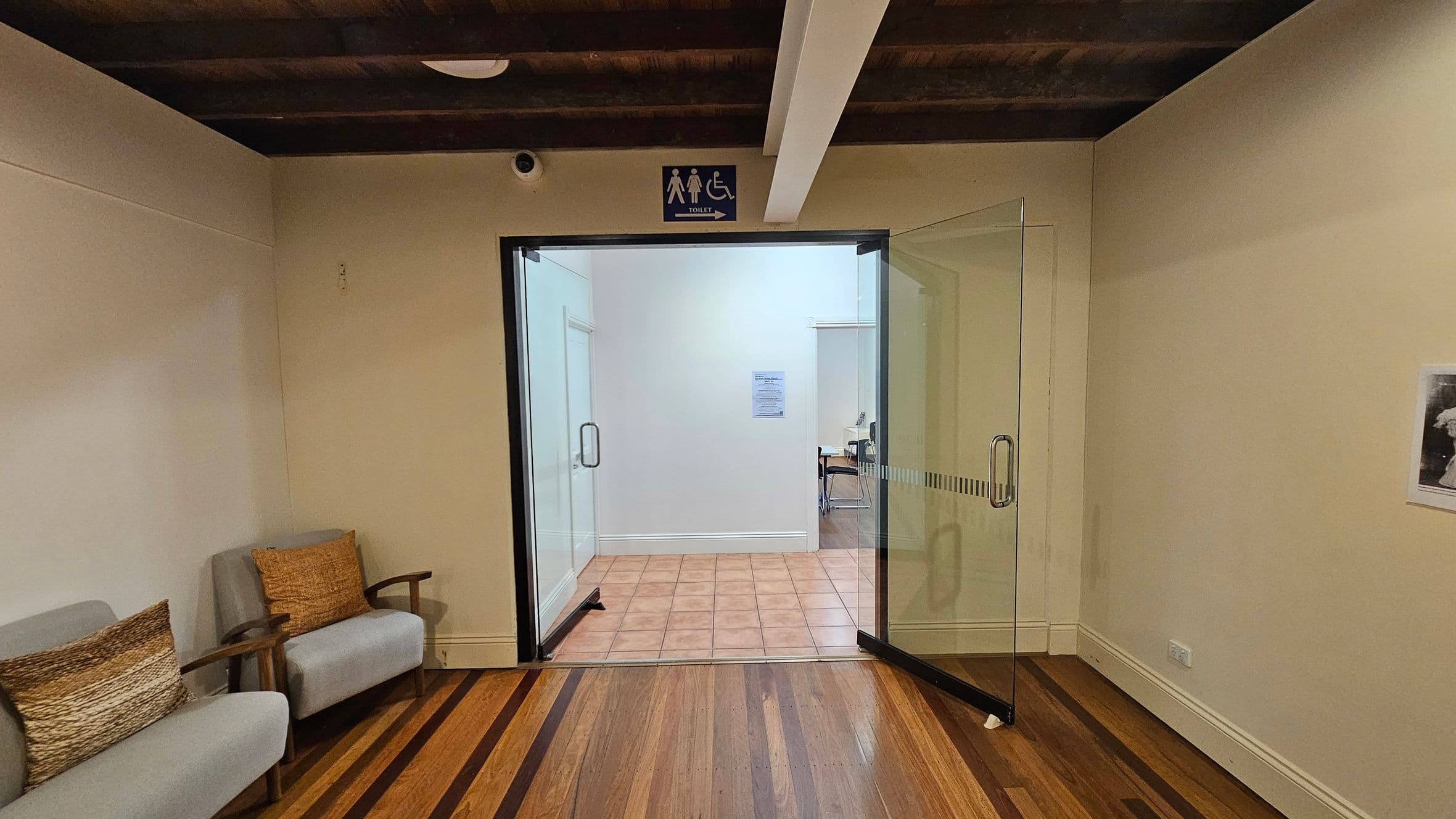 A room with wooden flooring and a wooden beam ceiling. On the left, there are two grey armchairs with brown cushions. In the centre, a glass door leads to another room with tiled flooring. Above the door, a sign indicates a toilet with male, female, and accessible symbols. On the right wall, a small framed picture is visible.