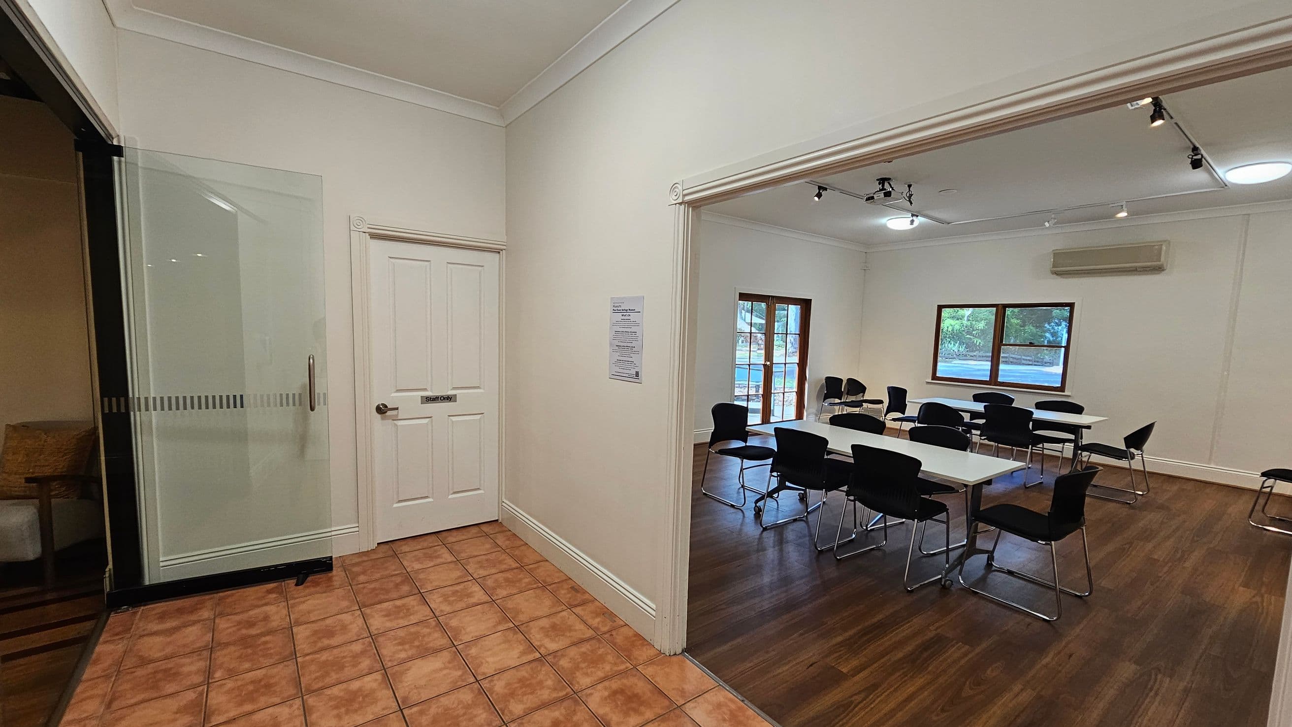 A room with a tiled floor in the foreground and a wooden floor in the background. On the left, there's a glass door with a handle, and a white door labelled "Staff Only". To the right, an open doorway leads to a room with several tables and black chairs. The room has two large windows and an air conditioning unit on the wall. The ceiling features recessed lighting and track lights.