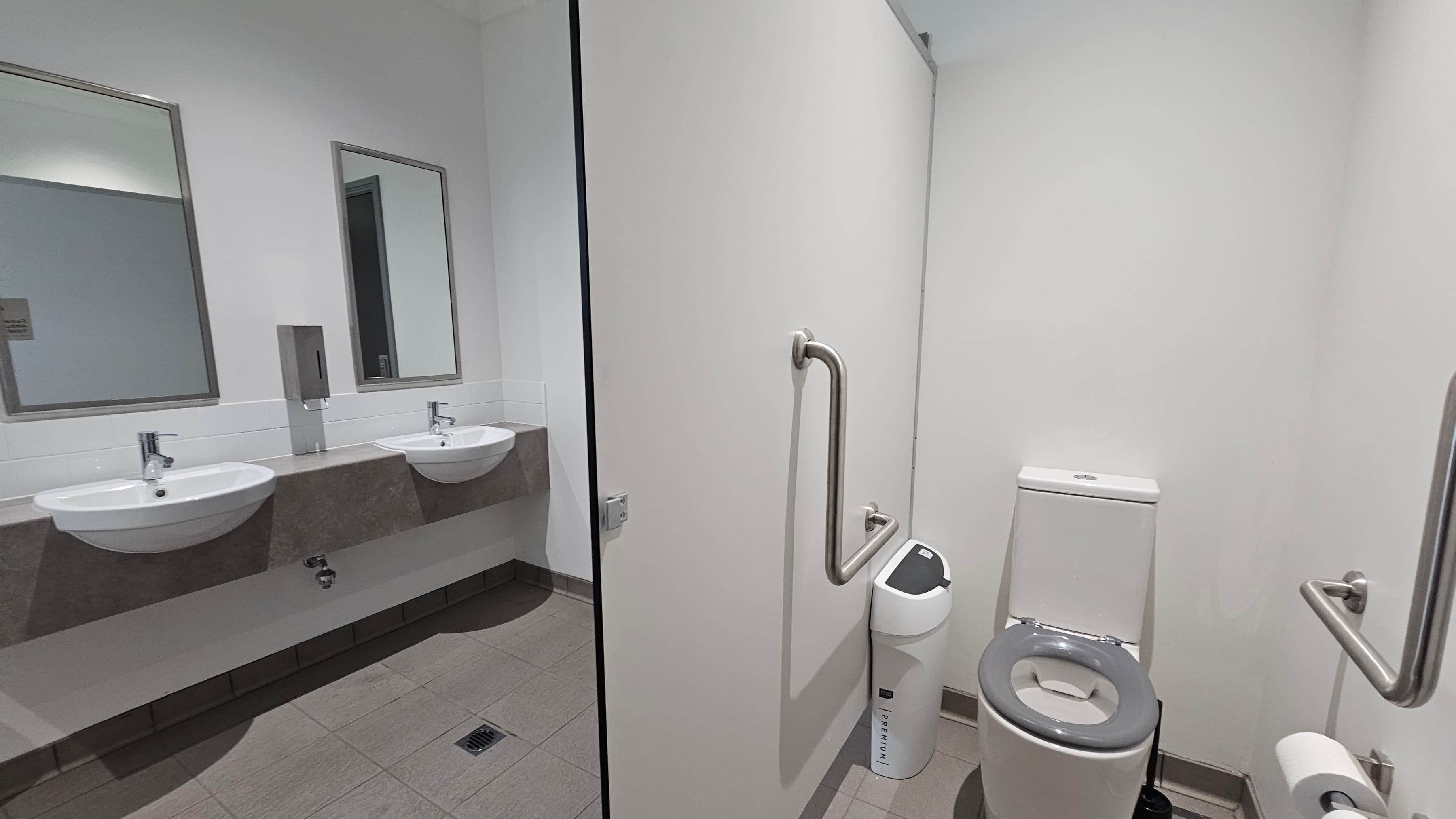 On the left, there are two white sinks with chrome taps set into a grey countertop, each with a rectangular mirror above. A soap dispenser is mounted between the mirrors. On the right, a white toilet with a grey seat is positioned next to a sanitary bin. A metal grab rail is attached to the wall beside the toilet. The floor is tiled in grey, and the walls are plain white.