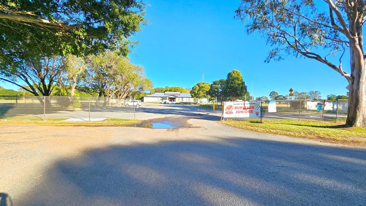 A paved road leads to a fenced area with a sign reading "Burleigh Bears Juniors" on the right. Behind the fence, a white building is visible, surrounded by trees. A white vehicle is parked to the left of the building. The foreground shows a shadowed road with a small puddle. Trees line both sides, with a large tree on the right and several on the left, casting shadows on the ground.
