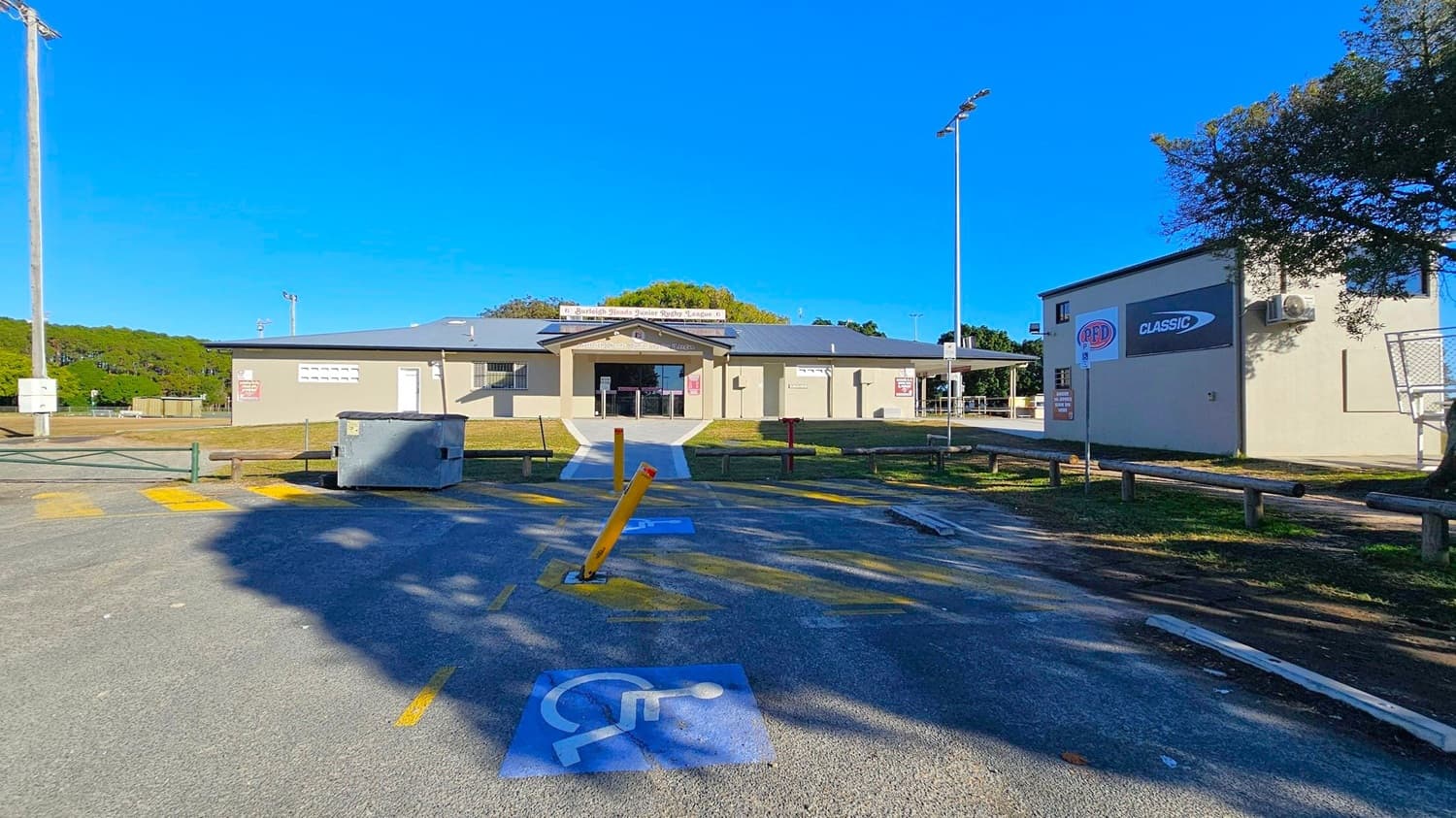 A single-storey building with a grey roof and beige walls is in the centre, featuring a sign above the entrance. In the foreground, a car park with a marked disabled parking space and a yellow bollard is visible. To the right, a tree provides shade, and a building with signage is adjacent. On the left, a grassy area and a utility pole are present. The background includes more trees and a clear blue sky.