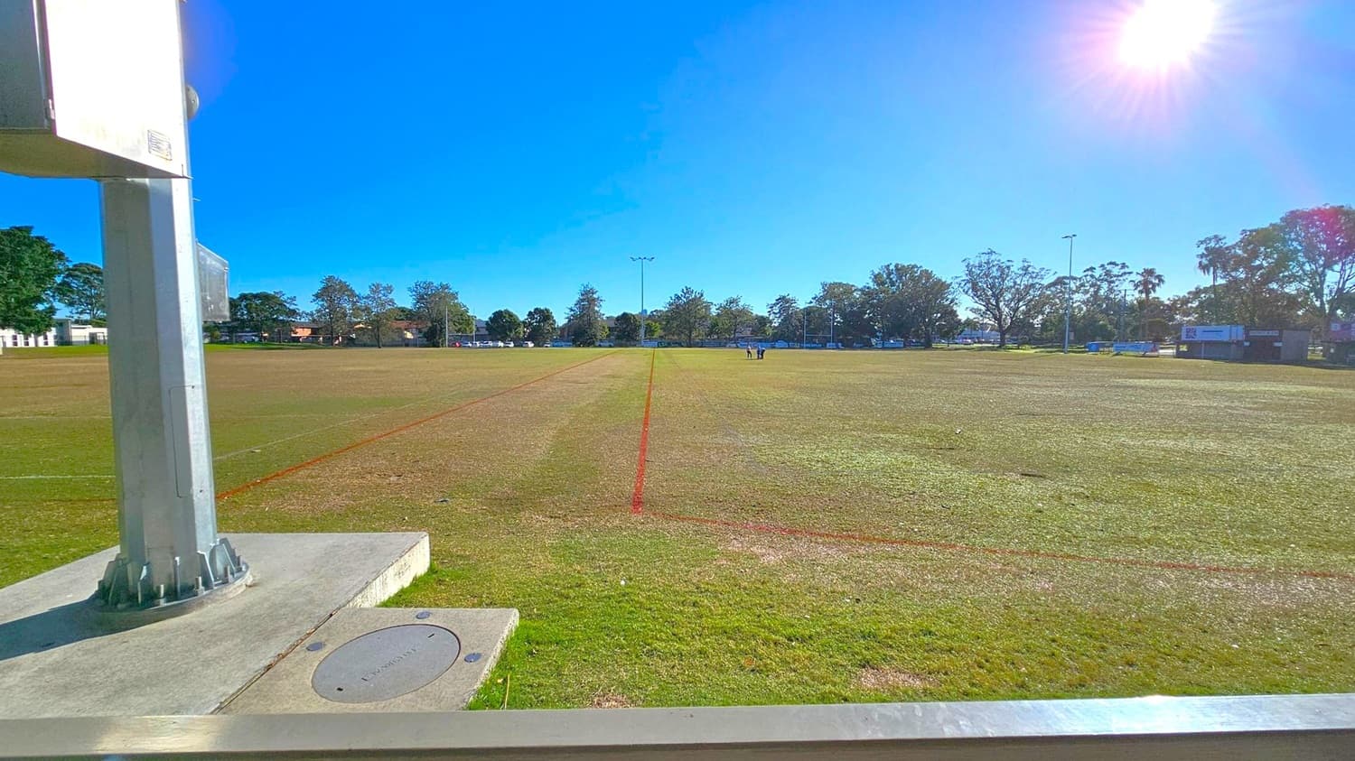 A sports field with a mix of green and brown grass, marked with red lines. In the foreground, a large metal post is mounted on a concrete base to the left. In the background, trees line the perimeter of the field, and a few people are visible in the distance. The sky is clear and blue, with the sun shining brightly in the top right corner.