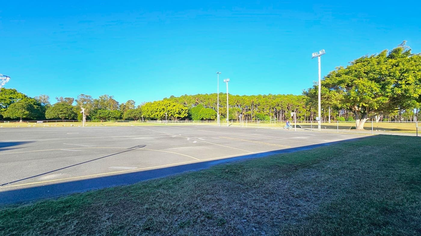 A large, open asphalt court with faint yellow lines is in the foreground, surrounded by a grassy area. On the left, a basketball hoop is partially visible. In the background, there are tall trees and a few scattered smaller trees. Two tall floodlights are positioned near the centre and right side. A person on a bicycle is near the right, close to a large tree. The sky is clear and blue.