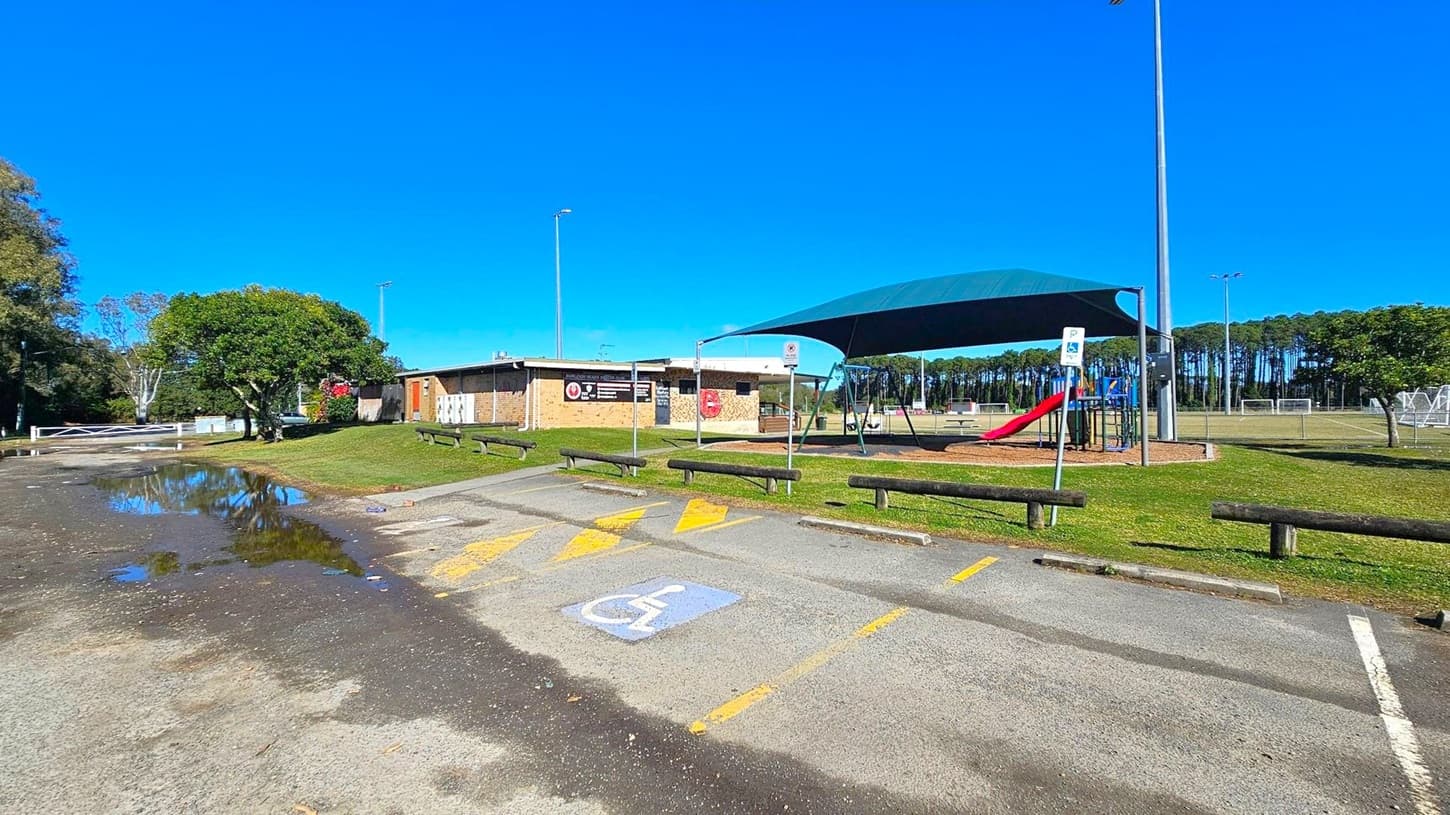 A playground with a blue shade cover and a red slide is on the right. Adjacent to it is a grassy area with wooden benches. In the centre, a brick building with signs is visible. A paved path with a disabled parking space marked in blue and yellow is in the foreground. On the left, a tree and a puddle are near a road. In the background, there are tall trees and a sports field with goalposts.