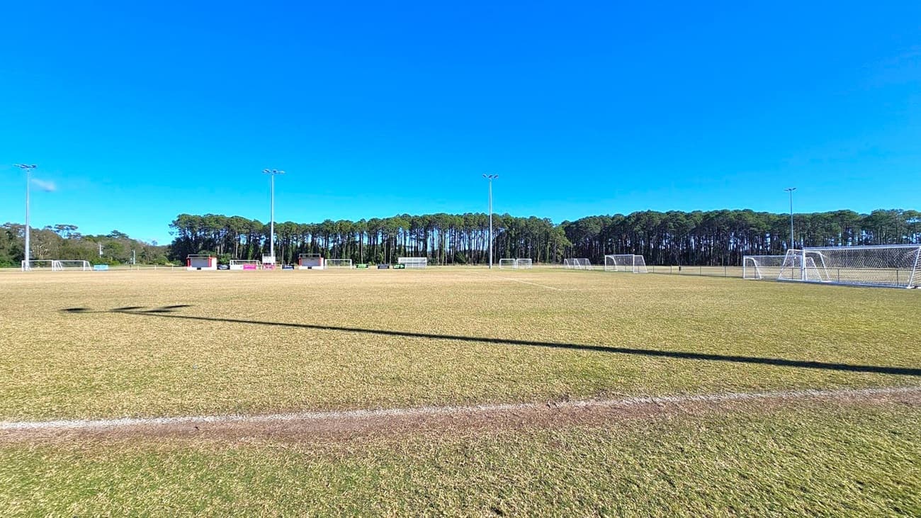 A football field with a dry, brownish grass surface. Multiple white goalposts are positioned across the field. Tall floodlights are evenly spaced along the perimeter. In the background, a dense line of tall trees stretches across the horizon. A few small structures, possibly for storage or seating, are visible near the trees. The sky is clear and blue.