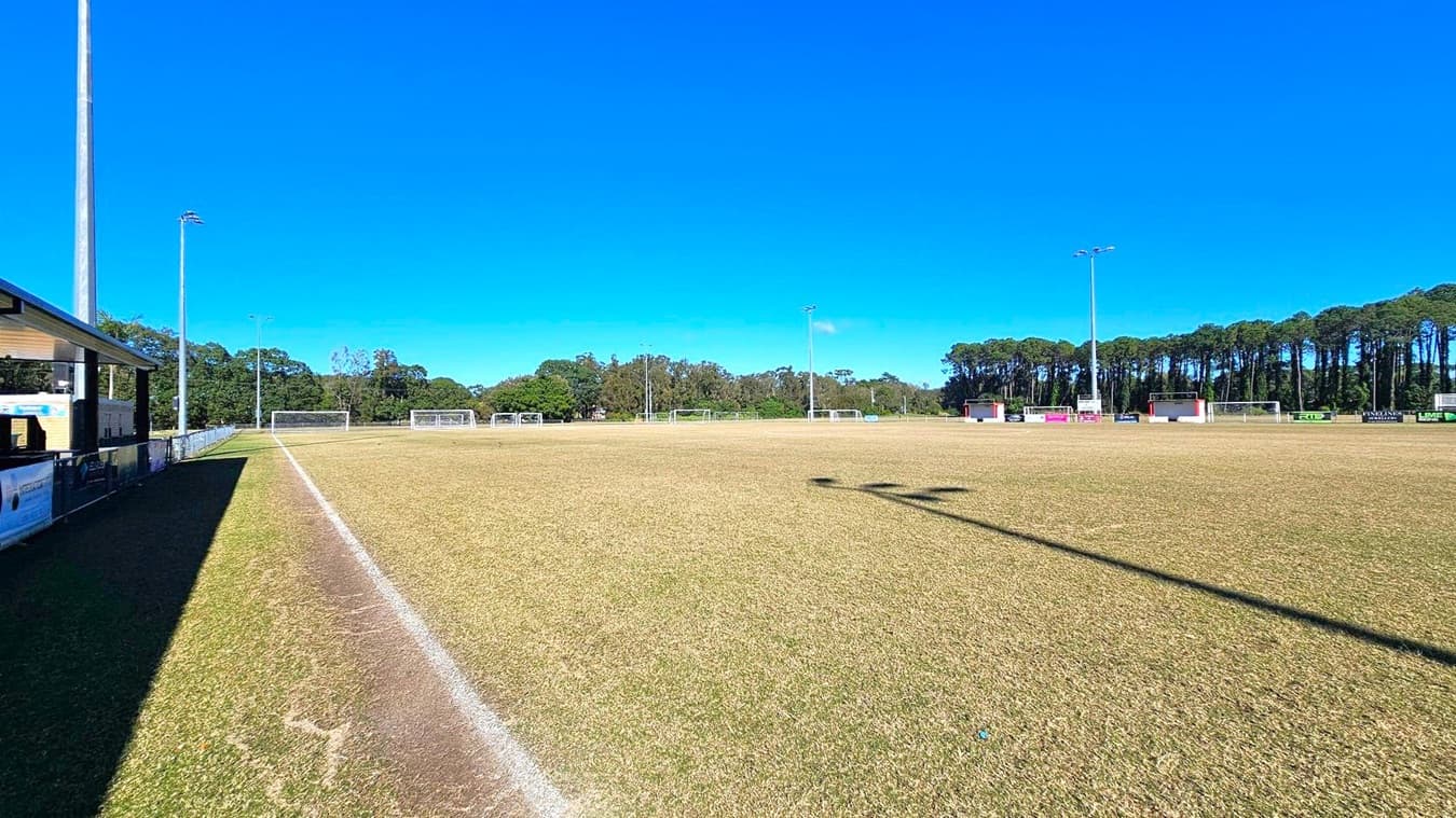 A football field with a dry, brownish grass surface. On the left, there is a building with a roof and a tall floodlight pole. The field is marked with white lines, and several goalposts are visible in the distance. In the background, there are trees lining the perimeter of the field. The shadows of the floodlights stretch across the grass.