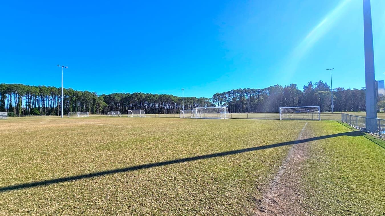 A football field with multiple white goalposts is visible. The grass is a mix of green and brown, indicating some wear. Tall floodlights are positioned around the field. In the background, there is a dense line of tall trees. A metal fence runs along the right side of the field. The sky is clear and blue, casting shadows on the ground.