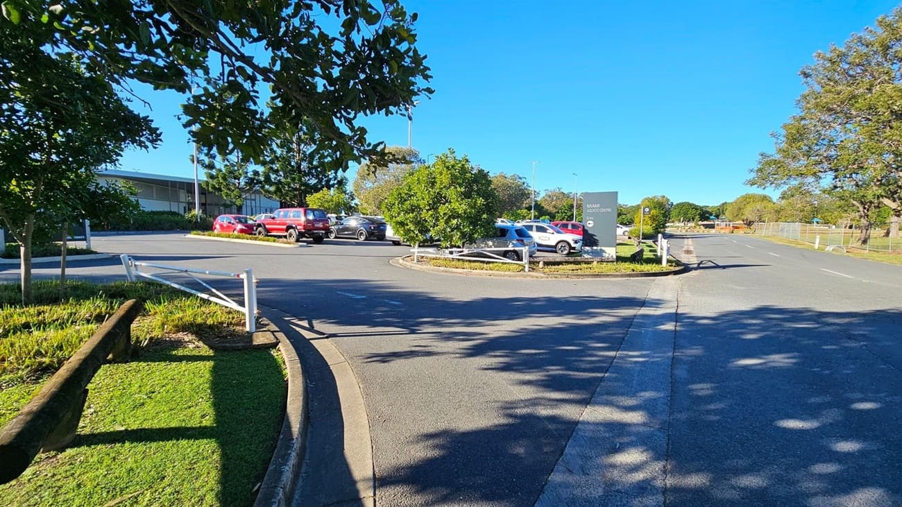 A car park with several vehicles, including red and white cars, is visible in the centre. On the left, there are trees and a building in the background. A sign on the right reads "MIAMI AQUATIC CENTRE." The road curves around the car park, with shadows from the trees cast on the asphalt. The area is surrounded by greenery, with more trees and a fence in the distance.