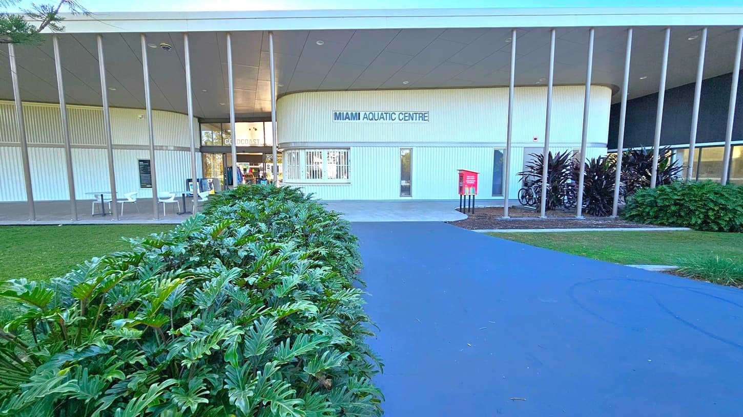 A modern building with a sign reading "Miami Aquatic Centre" on the front. The structure features vertical columns and a curved facade. On the left, there are white chairs and tables under a covered area. A path leads to the entrance, flanked by lush green plants. On the right, a red sign and bicycles are visible near more foliage. The ceiling is smooth and grey, with recessed lighting. The ground is paved with a dark surface.