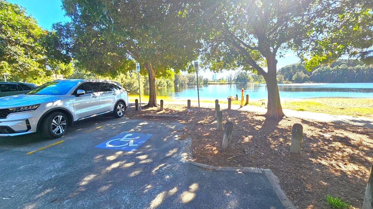 A white SUV is parked on the left in a designated disabled parking space, marked by a blue sign on the ground. The area is shaded by large trees, with sunlight filtering through the leaves. In the background, a calm body of water is visible, bordered by grassy areas and more trees. Wooden posts are scattered around the edge of the parking area, and a path runs alongside the water. The scene is peaceful, with a clear view of the water and distant greenery.