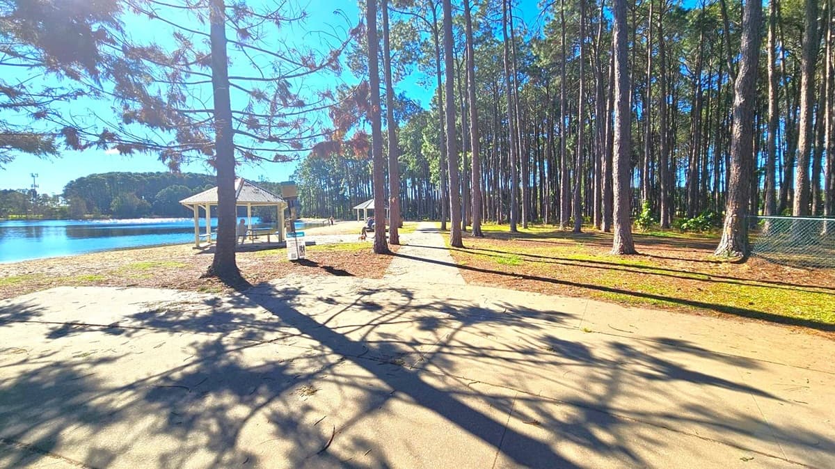 A paved path runs through a park with tall trees on the right casting long shadows. On the left, a small lake is visible with a gazebo near the water's edge. Another gazebo is further along the path. The ground is a mix of grass and dirt, and a chain-link fence is partially visible on the right. The scene is well-lit, suggesting a sunny day.