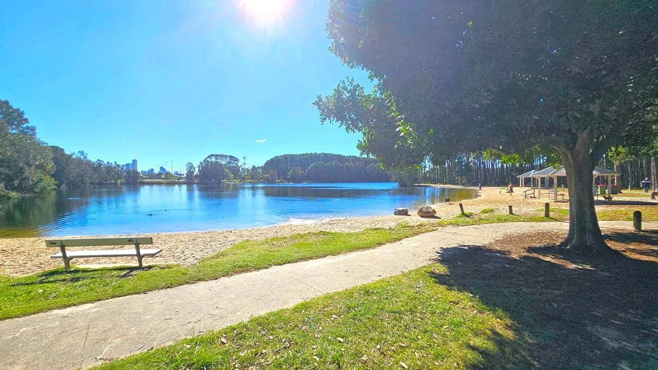 A serene lakeside scene with a bench on the left, facing a sandy beach and calm water. In the background, a city skyline is visible beyond the trees. On the right, a large tree provides shade over a grassy area, and a pathway leads to a couple of gazebos. People are scattered around, enjoying the sunny day. The foreground features a mix of grass and a paved path.