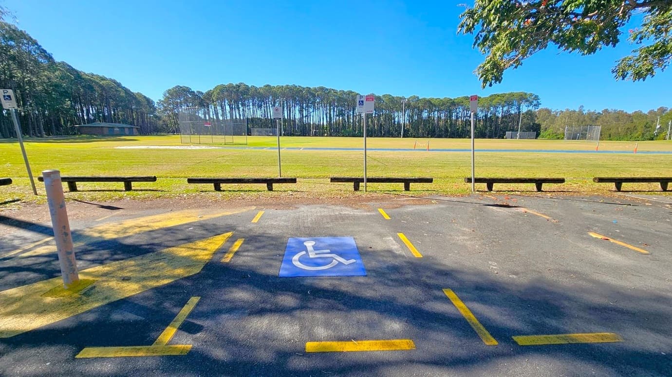 A parking area with a designated disabled parking space marked by a blue sign with a wheelchair symbol in the foreground. Yellow lines outline the space. In the background, there is a grassy field with a few sports nets and a line of tall trees. Wooden barriers separate the parking area from the field. A few signs are posted along the edge of the parking area. A tree with green leaves partially overhangs the scene from the right.