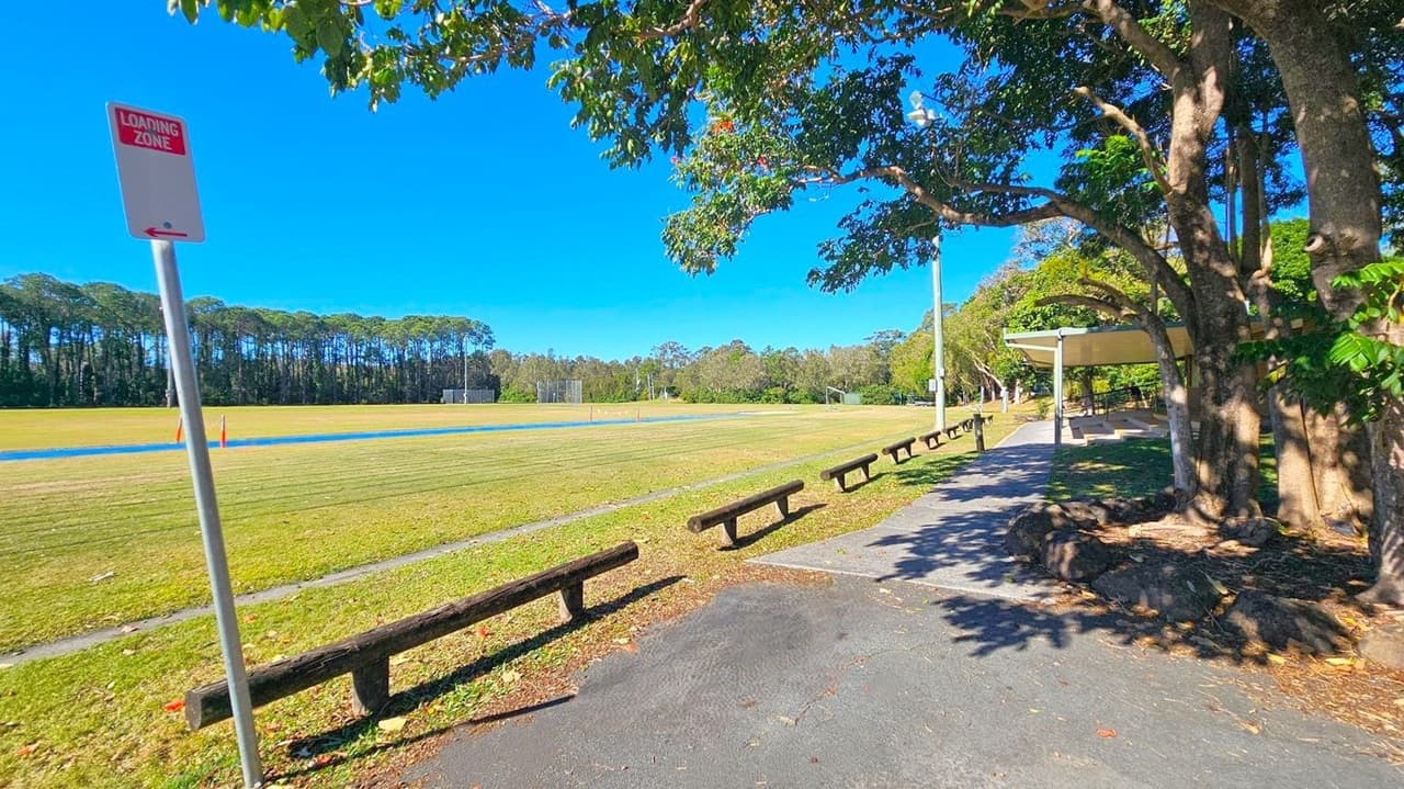 A grassy field stretches across the background, bordered by a line of tall trees. In the foreground, a paved path runs alongside the field, with wooden benches lining its edge. A "Loading Zone" sign is visible on the left. On the right, a large tree provides shade over the path, and a covered seating area is partially visible. The ground is scattered with fallen leaves, and the area is well-lit by sunlight.