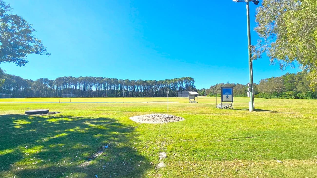 A grassy sports field with a sandy patch in the foreground. On the right, there's a tall light pole and a small structure with a sign. In the background, a line of tall trees borders the field. A small shelter with a roof is visible near the centre. Shadows from trees are cast on the grass, and the area is open and spacious.