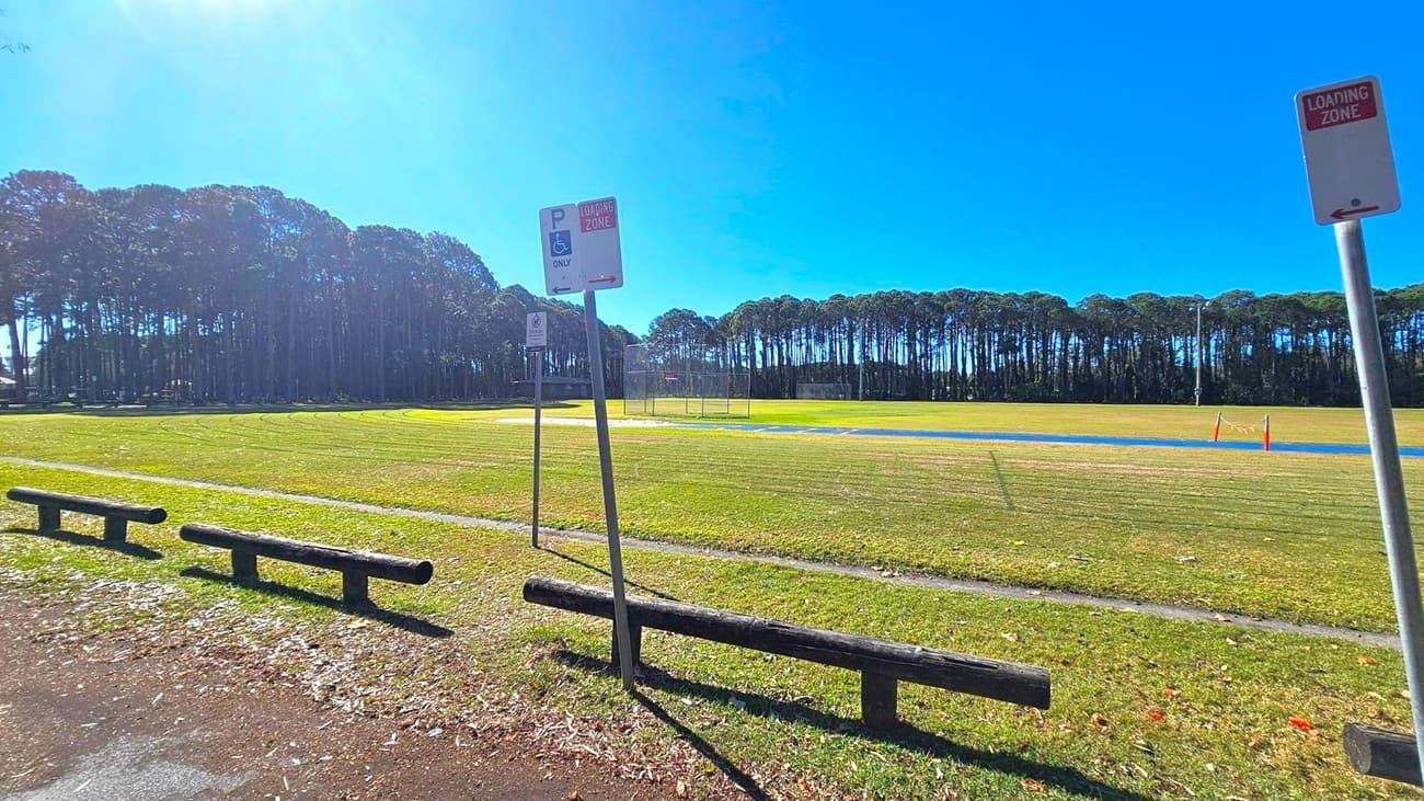 A grassy field with a line of tall trees in the background. In the foreground, there are wooden barriers and a paved path. Several signs are visible, including a "Loading Zone" sign and a parking sign with a wheelchair symbol. The field has a blue track and some orange cones. The sky is clear and blue.