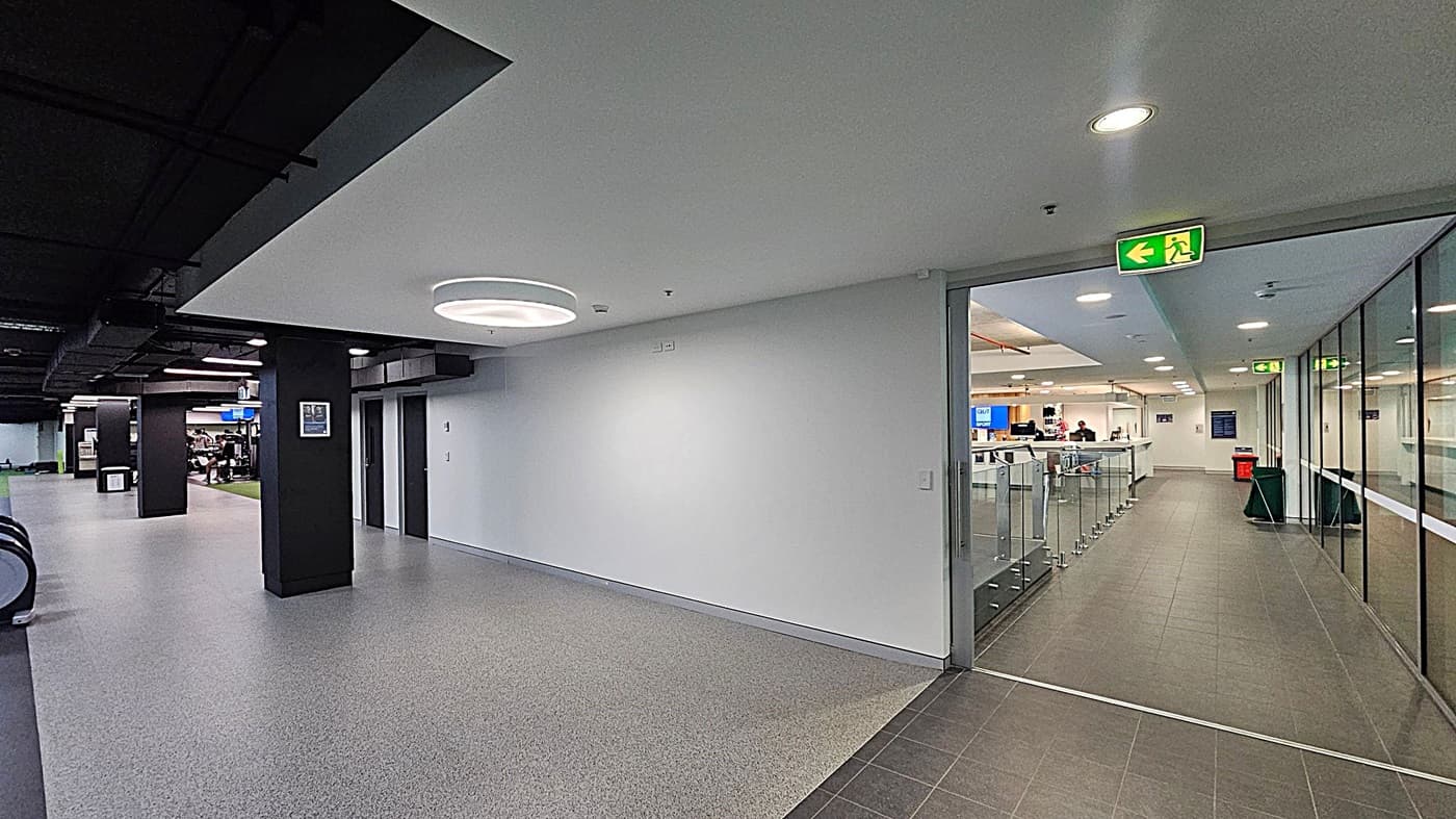A modern indoor space with a grey speckled floor. On the left, there are escalators and a gym area with exercise equipment. The ceiling is black with recessed lighting. On the right, a glass-walled corridor leads to a reception area with a counter and people behind it. An illuminated green exit sign is above the glass doors. The walls are white, and there are black columns throughout the space.