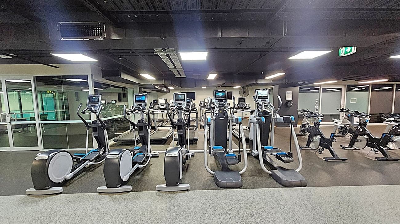A gym with a row of elliptical machines and treadmills in the centre. To the right, there are several stationary bikes. The floor is covered with a speckled grey and green carpet. The ceiling is black with rectangular lights. On the left, there is a glass wall with a door leading to another room. An exit sign is visible on the ceiling to the right.