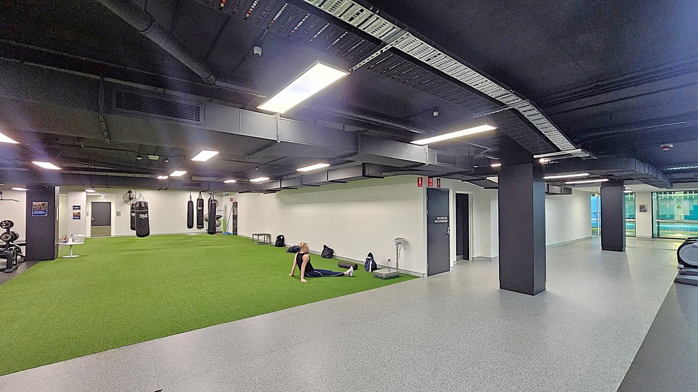 A gym interior with a large area of green artificial turf on the left, where a person is sitting on the floor. Several punching bags hang from the ceiling. On the right, there is a grey speckled floor with black pillars and a treadmill. The ceiling is black with exposed ductwork and bright rectangular lights. In the background, there are doors and a window showing an outdoor view.