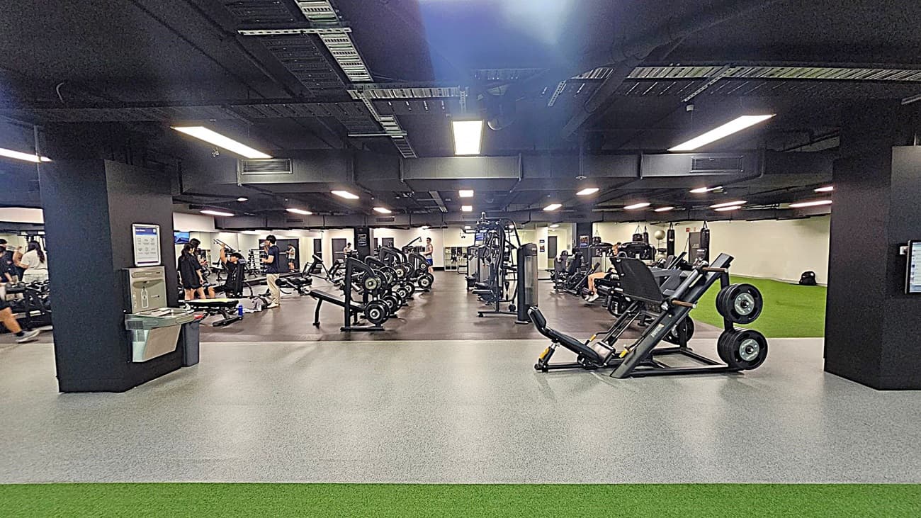 A spacious gym with various exercise equipment. On the left, there are people using weight machines and benches. In the centre, there are rows of weightlifting equipment and machines. On the right, a person is walking on a green artificial turf area. The floor is a mix of grey and green surfaces, and the ceiling is black with exposed ductwork and bright overhead lights.