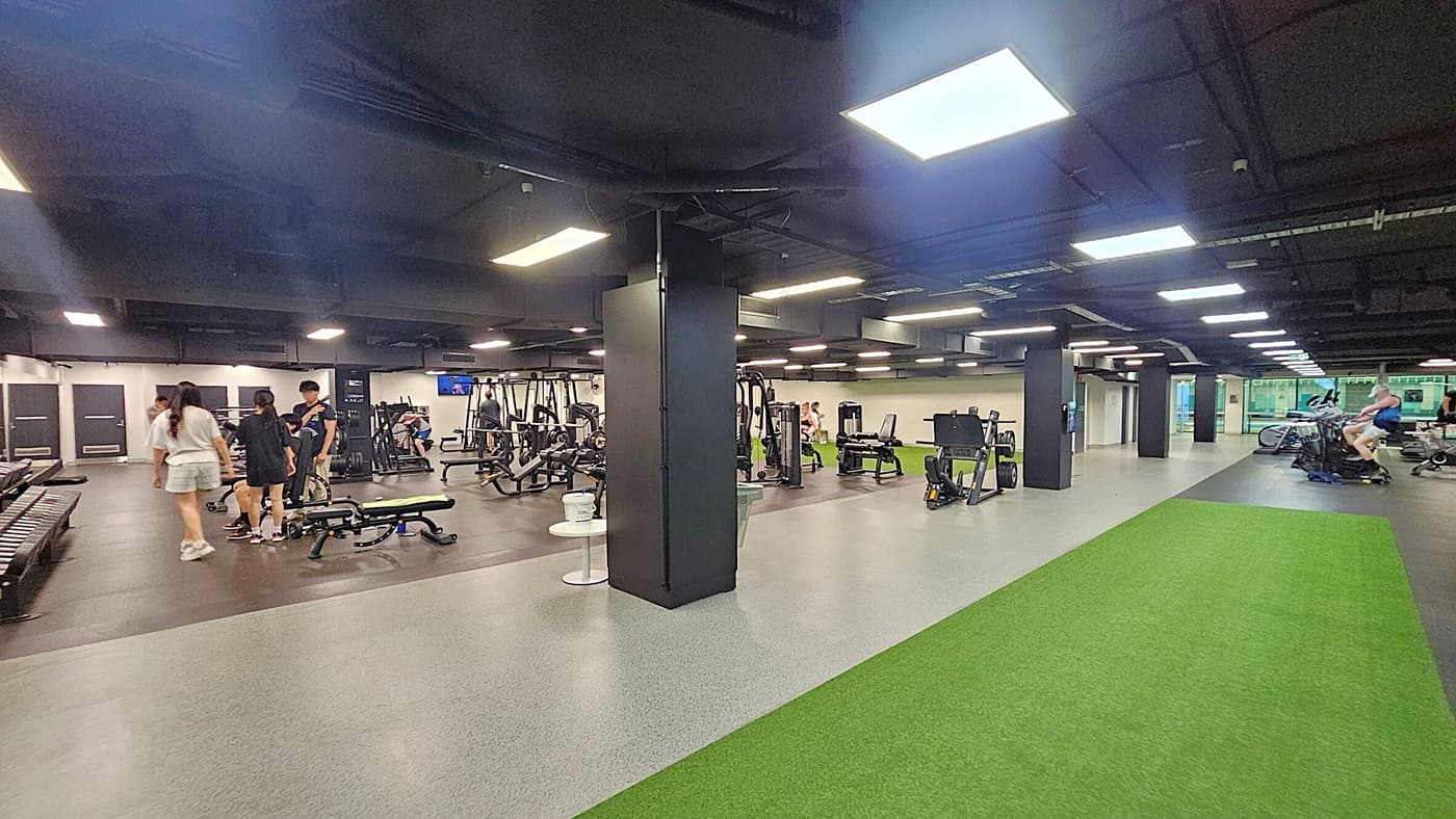 A spacious gym interior with various exercise equipment. On the left, several people are gathered near weight benches and machines. In the centre, there are more weight machines and a black pillar. On the right, a green turf strip runs along the floor, with cardio machines like stationary bikes nearby. The ceiling is dark with bright rectangular lights, and the floor is a mix of grey and green surfaces.