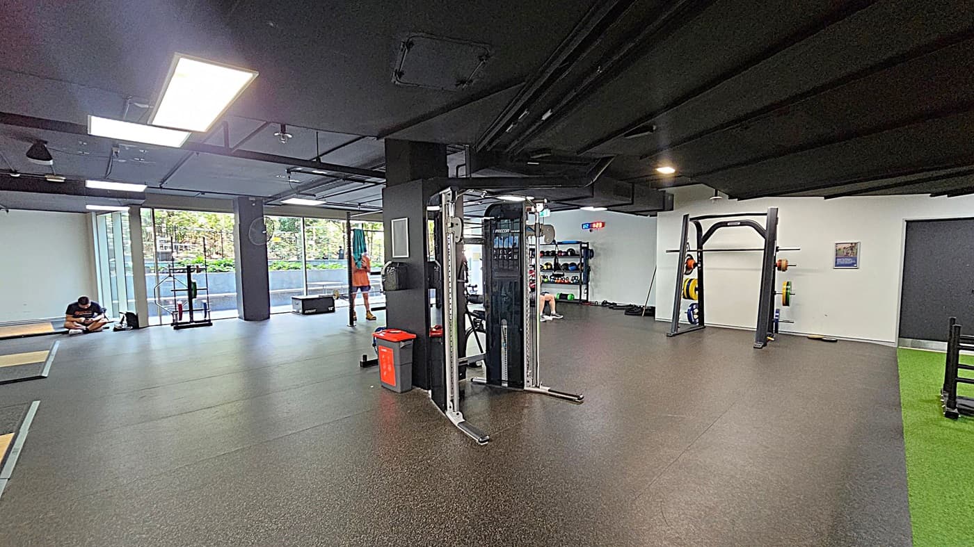 A spacious gym with a dark ceiling and rubber flooring. On the left, a person sits on the floor near large windows, with gym equipment nearby. In the centre, a multi-functional exercise machine and a red bin are visible. On the right, there is a squat rack with weights and a green turf area. The back wall has shelves with kettlebells and other equipment. Bright overhead lights illuminate the space.