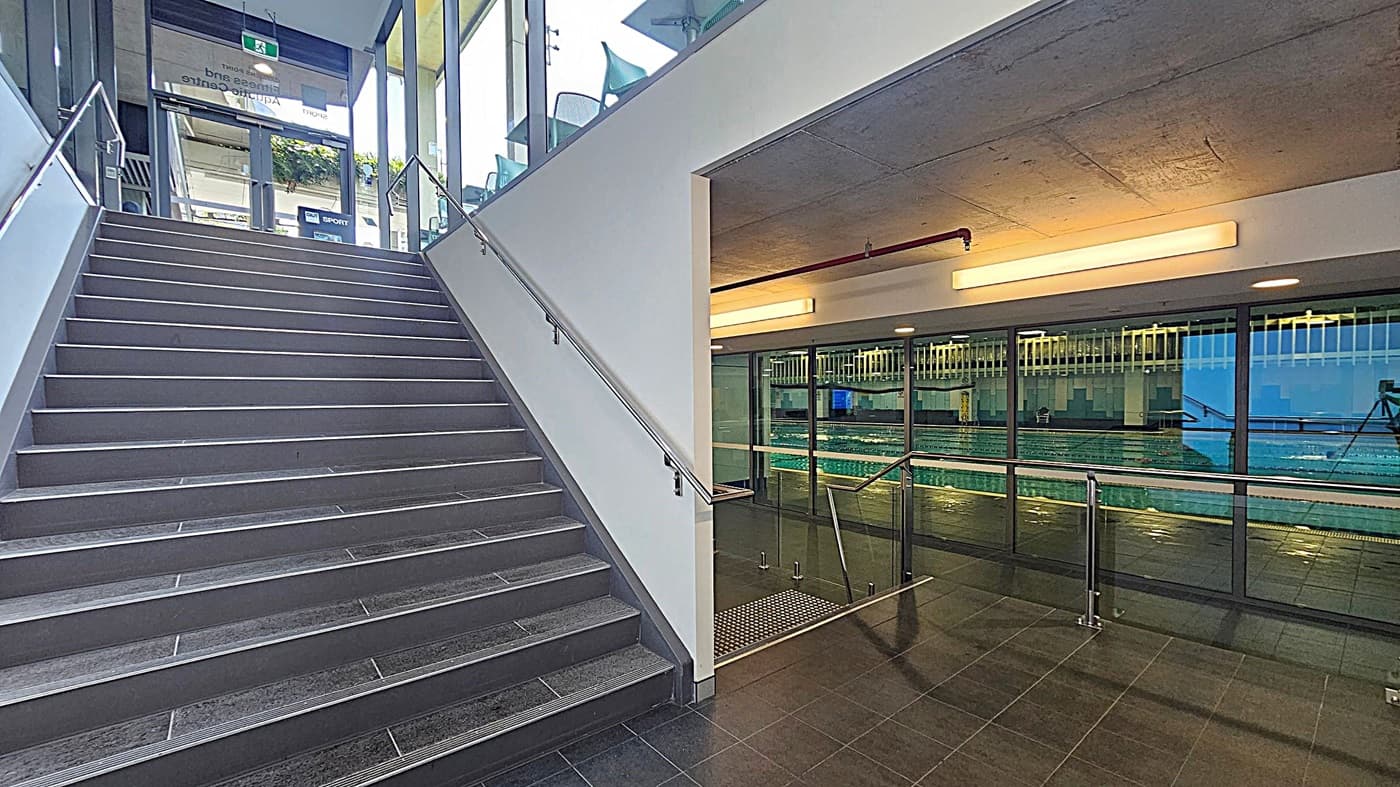 A modern indoor space with a staircase on the left, featuring grey steps and metal railings. To the right, there is a glass wall with a view of an indoor swimming pool. The ceiling is concrete with long, rectangular lights. The floor is tiled in dark grey. The area is well-lit, and there are glass doors at the top of the stairs leading outside.
