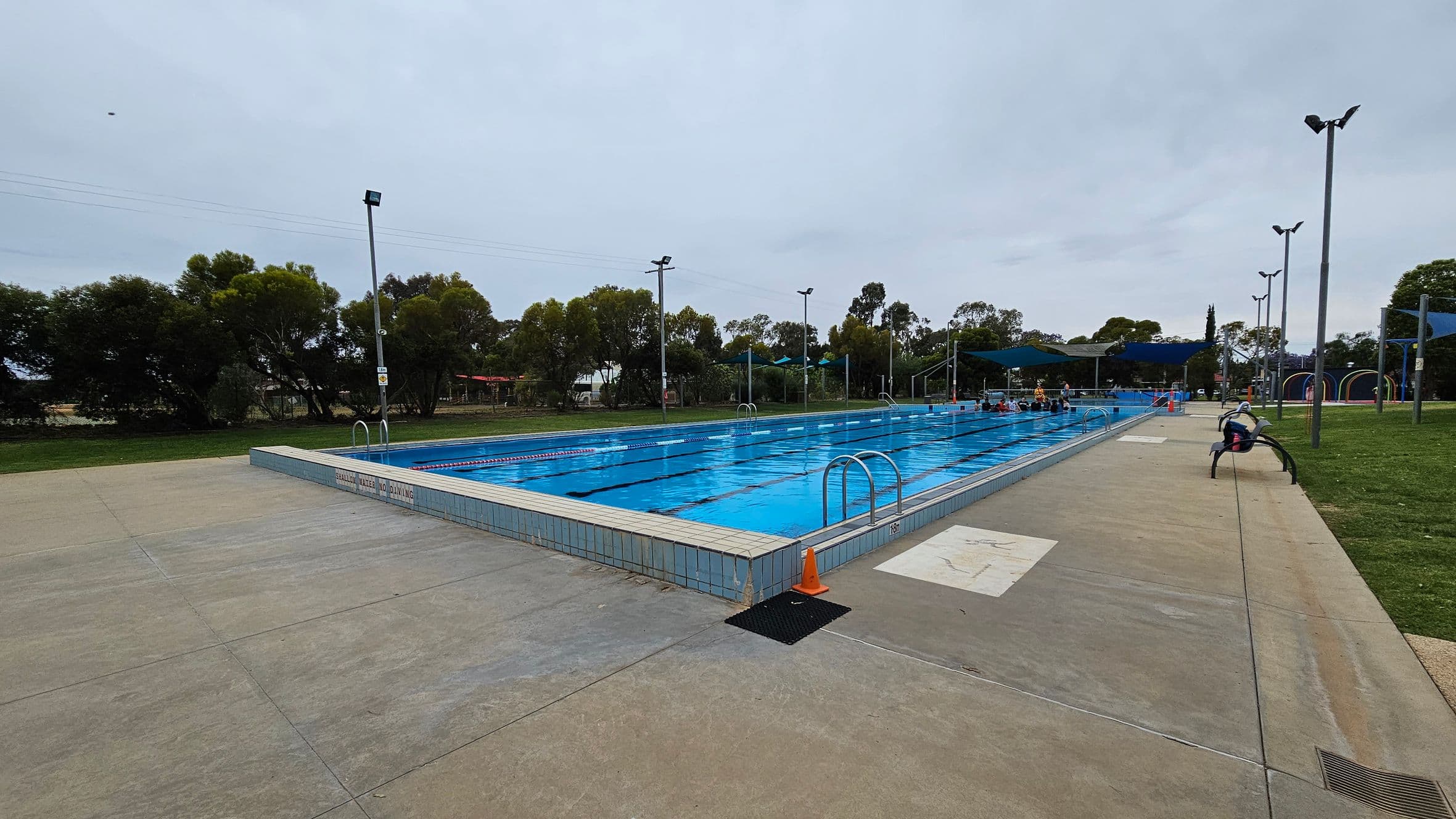 An outdoor swimming pool with clear blue water is in the centre, surrounded by a tiled deck. On the right, a person sits on a bench near the pool's edge. Several tall lights line the poolside. In the background, there are trees and some shaded areas with blue canopies. The ground is a mix of green and beige concrete. A small orange cone is placed near the pool's corner.