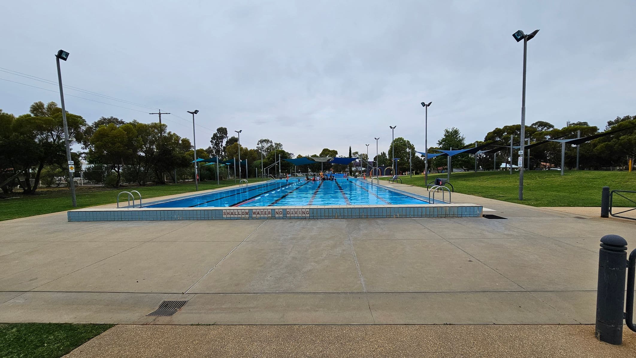 An outdoor swimming pool with clear blue water is in the centre, surrounded by a paved area. Signs on the pool edge read "SHALLOW WATER NO DIVING." Several tall lights are positioned around the pool. Green grass and trees are visible in the background, with some shade structures on either side. The sky is overcast.