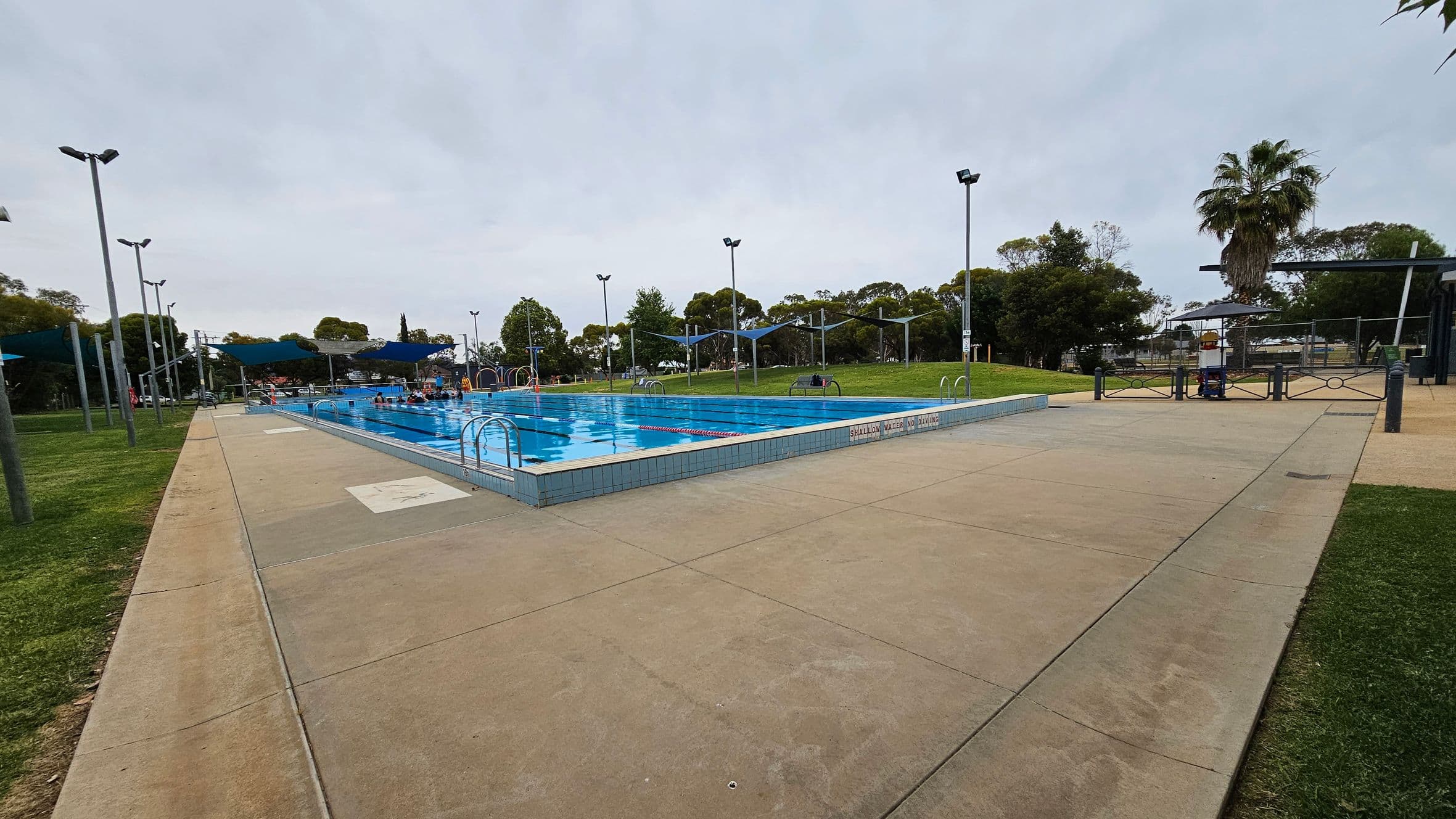 An outdoor swimming pool is surrounded by a concrete deck. On the left, there are several tall light poles and a grassy area. The pool is in the centre, with people swimming and blue shade sails overhead. On the right, a palm tree and more greenery are visible. A fence and gate are at the far end of the pool area. The pool has a sign indicating "shallow water."