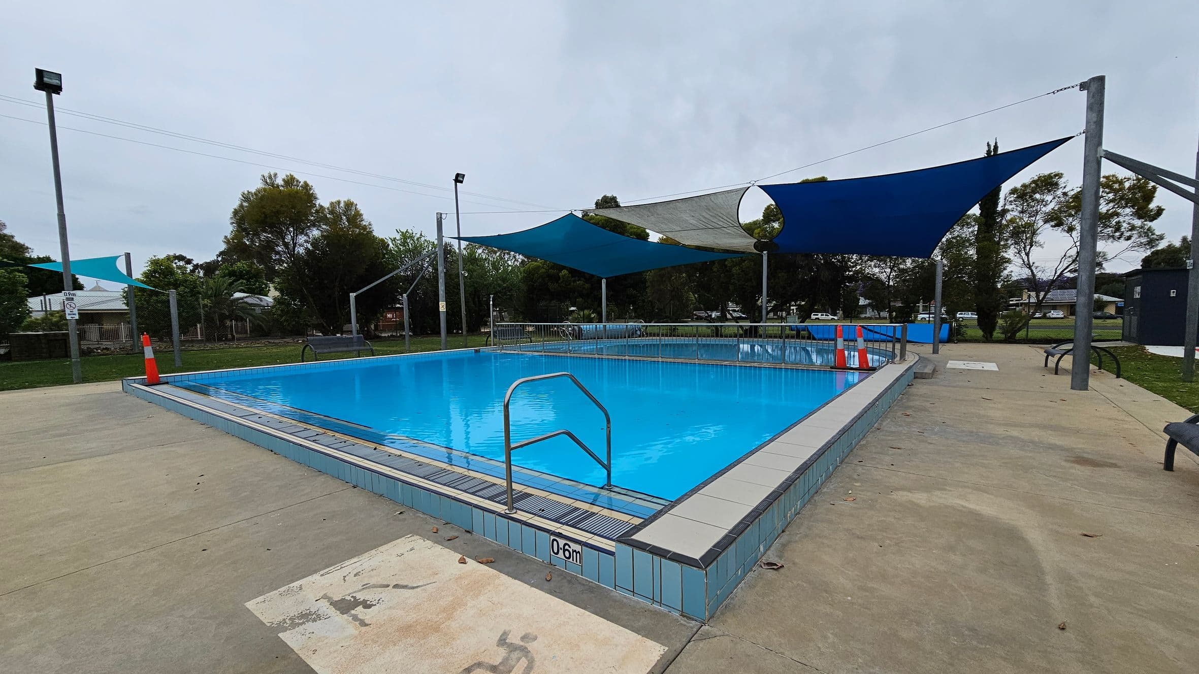 An outdoor swimming pool with clear blue water is surrounded by a tiled edge. A metal handrail descends into the pool. Overhead, several triangular shade sails in blue and grey are suspended. The pool area is bordered by a concrete surface with a traffic cone near the edge. In the background, there are trees and a grassy area, with a few benches and a fence visible.