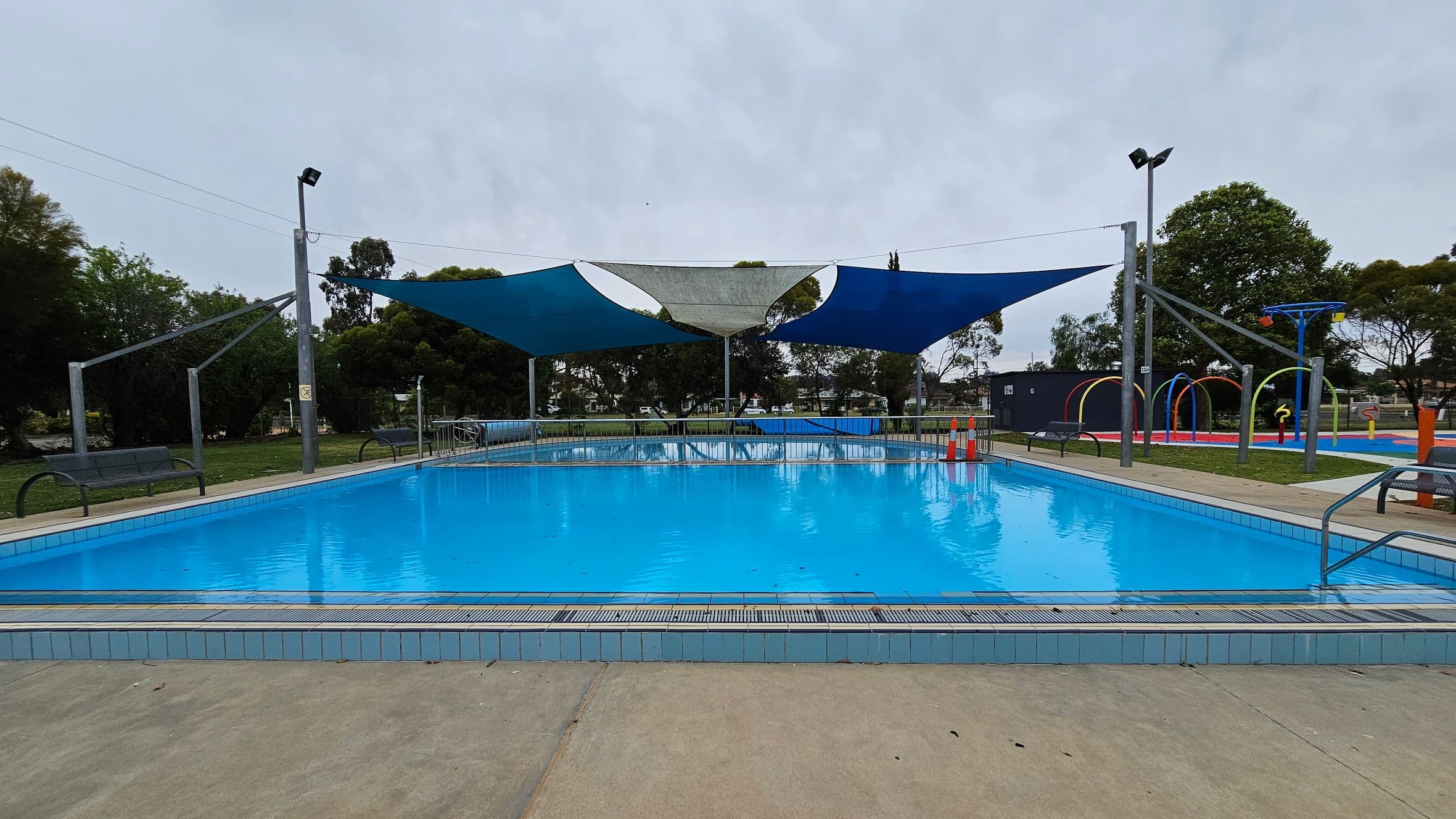 A rectangular outdoor swimming pool with clear blue water is in the centre. Overhead, there are triangular shade sails in blue and grey. Surrounding the pool are metal benches and a concrete walkway. In the background, there are trees and a colourful water play area with arches and water features. Orange traffic cones are placed near the pool's edge.