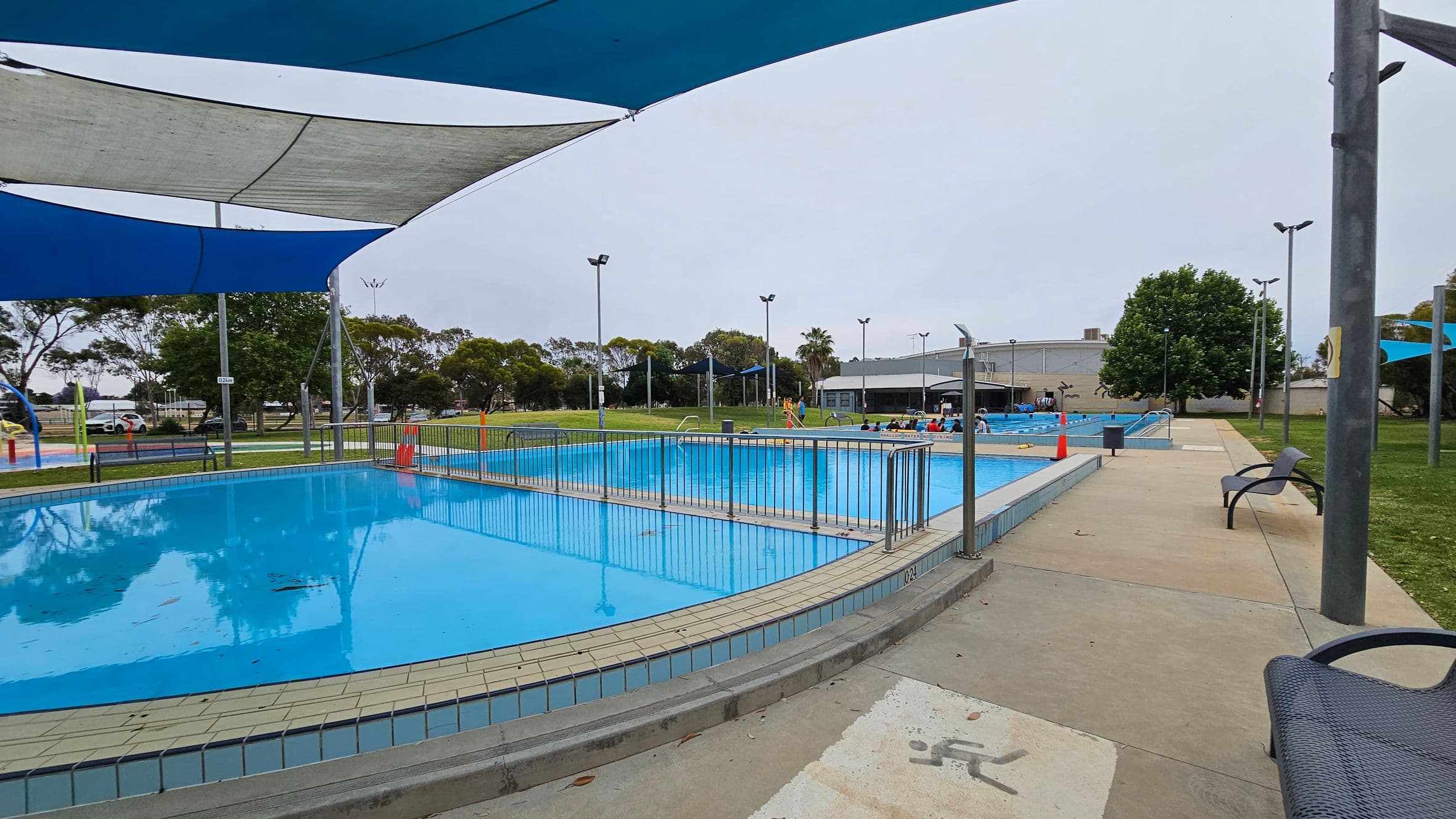 A swimming pool area with two pools separated by a metal fence. The left pool is covered by blue and grey shade sails. On the right, a paved walkway with a few chairs leads to a grassy area with trees. In the background, there are people near the second pool, a building, and more trees. The ground is a mix of concrete and grass.