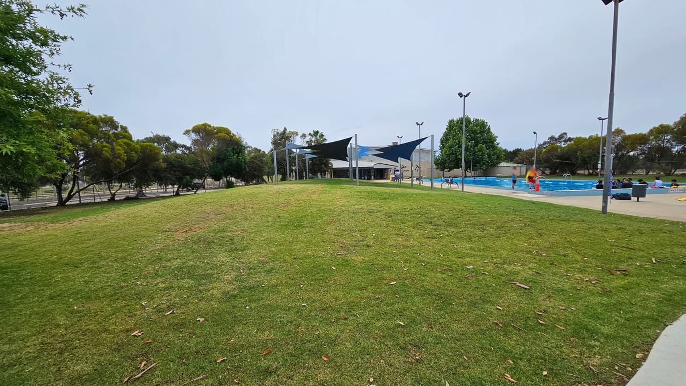 A grassy park area with scattered leaves in the foreground. In the background, there is a building with a series of triangular shade sails. To the right, a swimming pool is visible with people around it, including a person in a red outfit. Trees line the left side, and there are tall light poles near the pool.