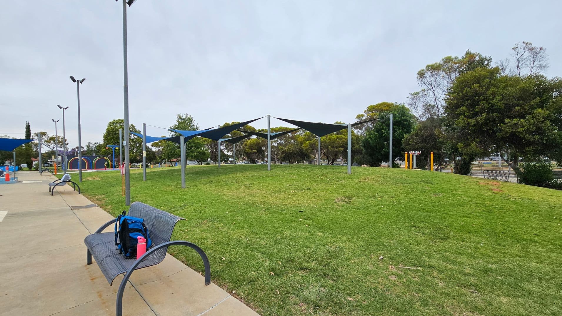 A park scene with a grassy area in the centre, featuring several triangular shade structures supported by metal poles. On the left, a paved path runs alongside the grass, with metal benches placed along it. One bench has a blue backpack and a pink bottle on it. In the background, there are trees and a playground with colourful arches. The area is well-lit with tall street lamps.