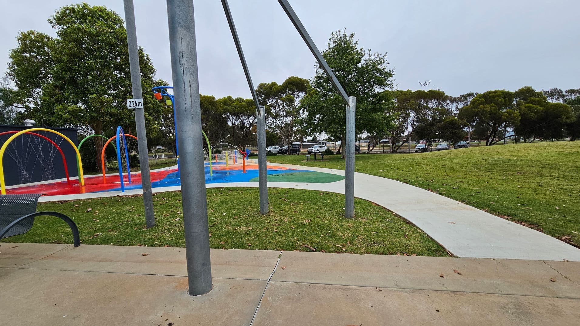 A park scene with a splash pad on the left featuring colourful arches and water play equipment. A sign on a pole reads "0.24m". A concrete path curves through the grassy area on the right. Trees line the background, and several cars are parked in the distance. A bench is partially visible in the foreground on the left, and the ground is a mix of grass and concrete.
