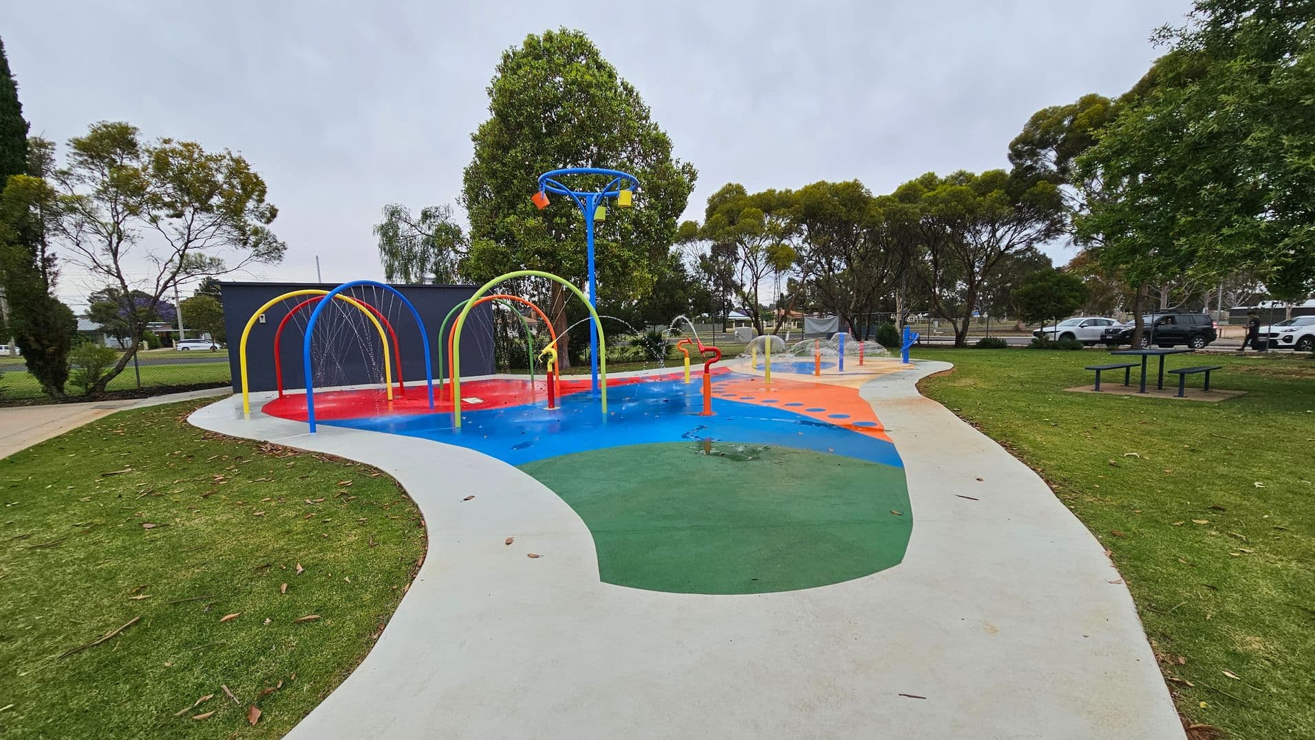 A colourful splash pad with red, blue, and green sections, featuring water arches and sprinklers. On the left, there are trees and a dark building. In the background, more trees and parked cars are visible. The foreground has a curved concrete path and grassy areas with scattered leaves. A picnic table is on the right side.