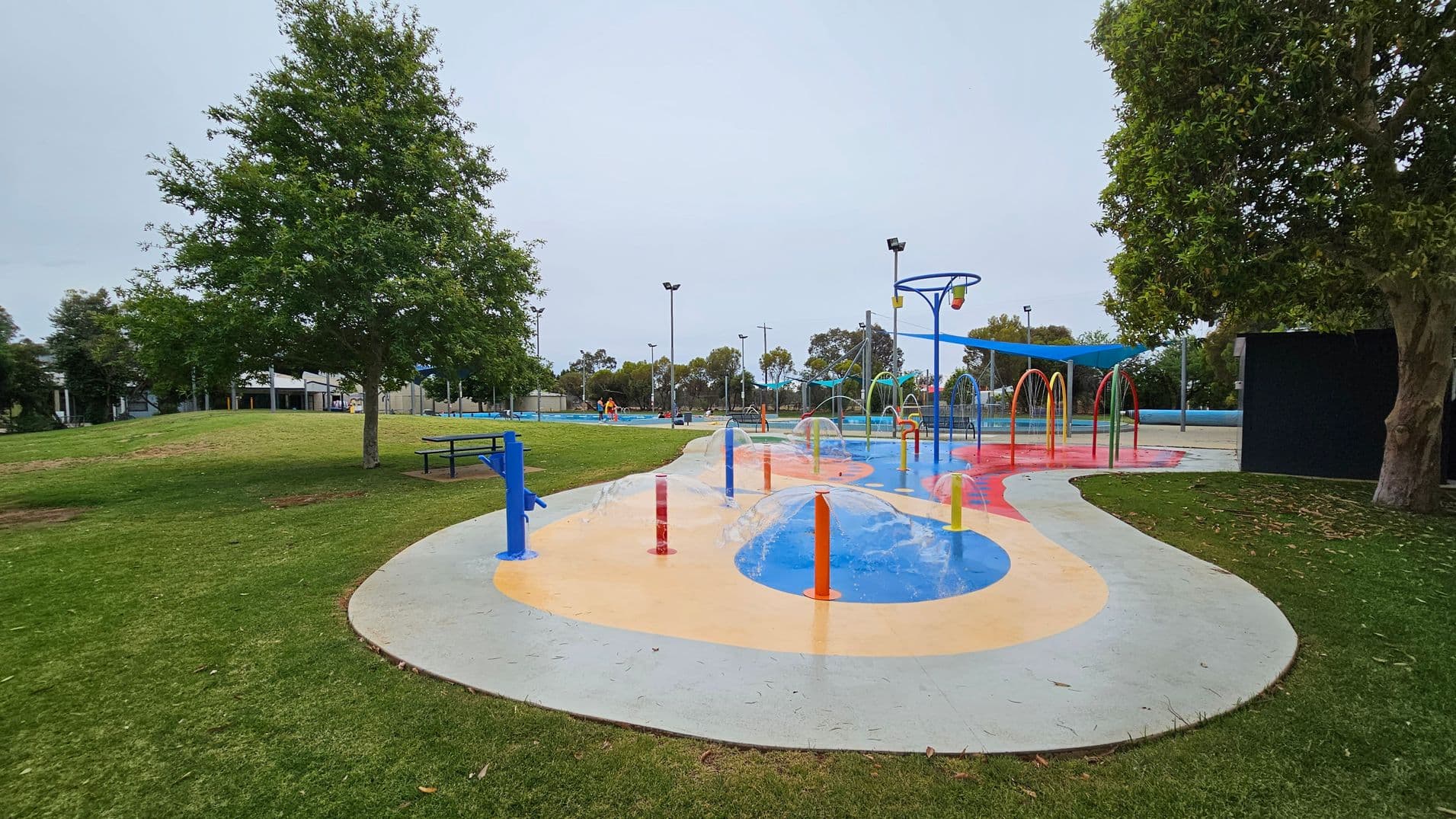 A colourful splash pad with water jets and hoops is in the centre, surrounded by a concrete path. On the left, there's a grassy area with a large tree and a picnic table. To the right, another tree provides shade. In the background, there are more trees, a pool, and a few people. The area is well-lit with tall lampposts.