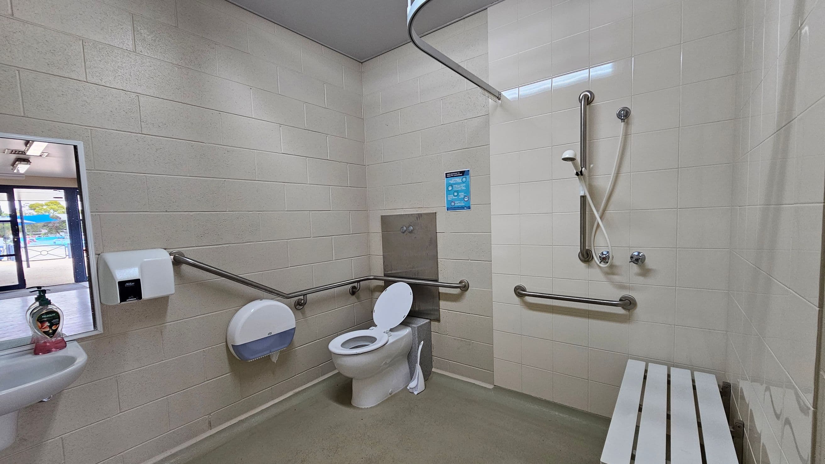 A bathroom with white tiled walls and a green floor. On the left, there's a small sink with a soap dispenser, a hand dryer, and a toilet paper dispenser. In the centre, a toilet with grab bars on both sides. On the right, a shower area with a handheld showerhead and additional grab bars. A white bench is attached to the wall. A sign is posted above the toilet. A window on the left shows an outdoor area with a pool.